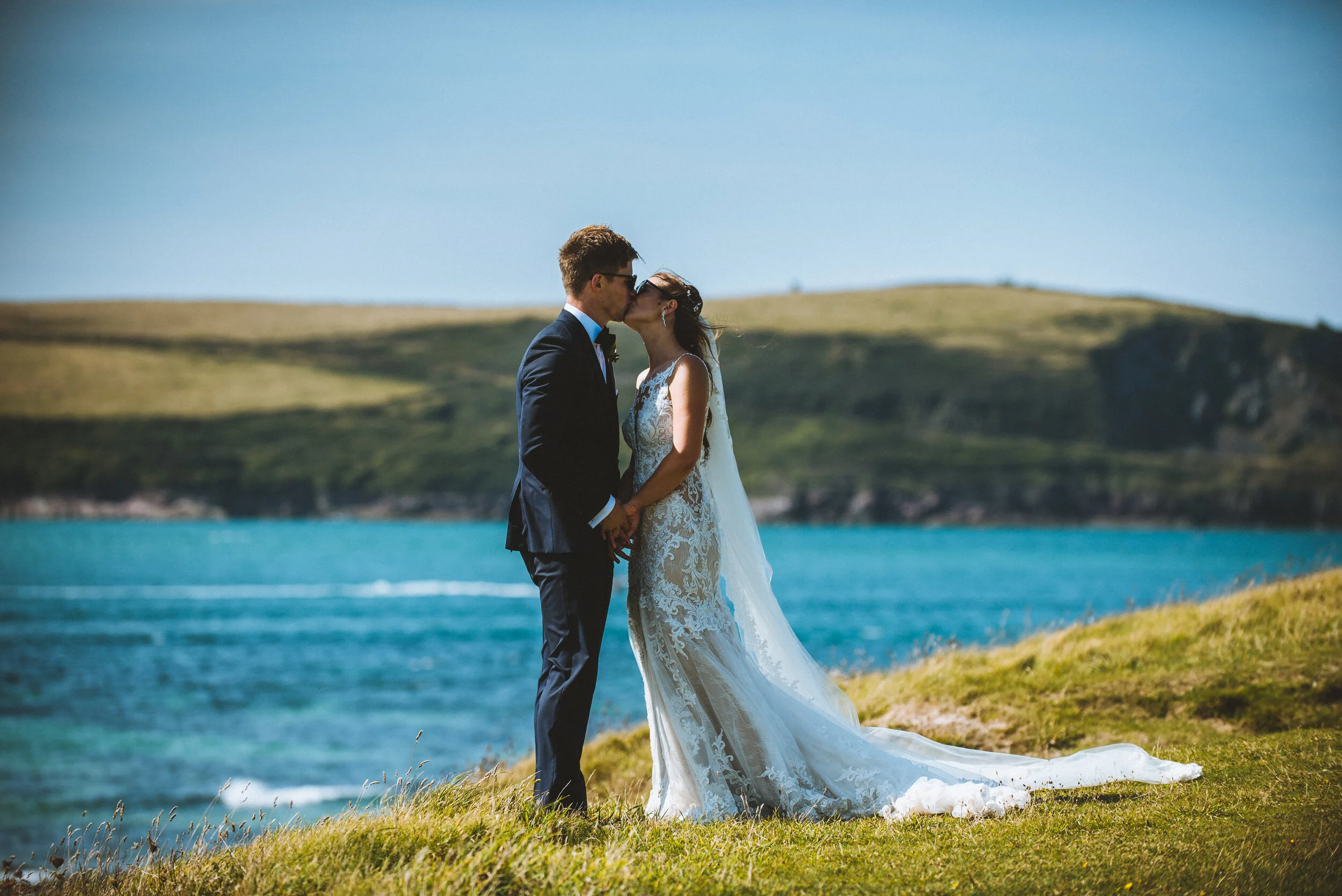 A bride and groom sharing a kiss by the water on a sunny day, with green hills in the background.