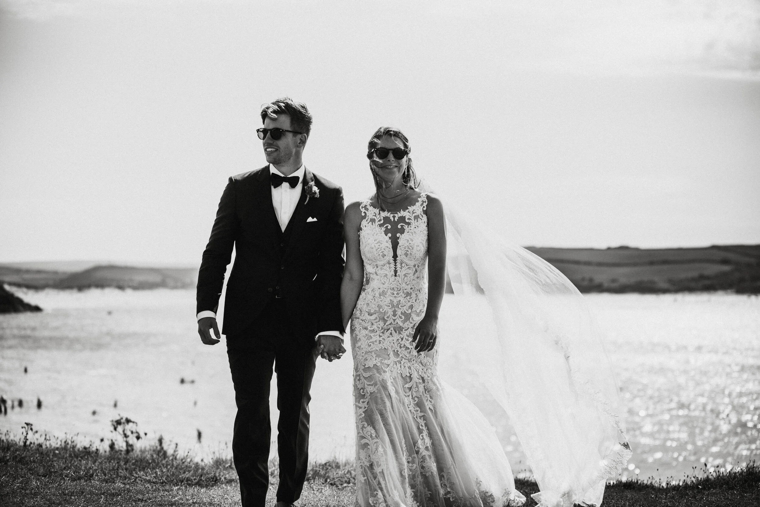 Isles of scilly photography. A black and white photo of a bride and groom holding hands, walking along a shoreline, both wearing sunglasses, with water and hilly terrain in the background.
