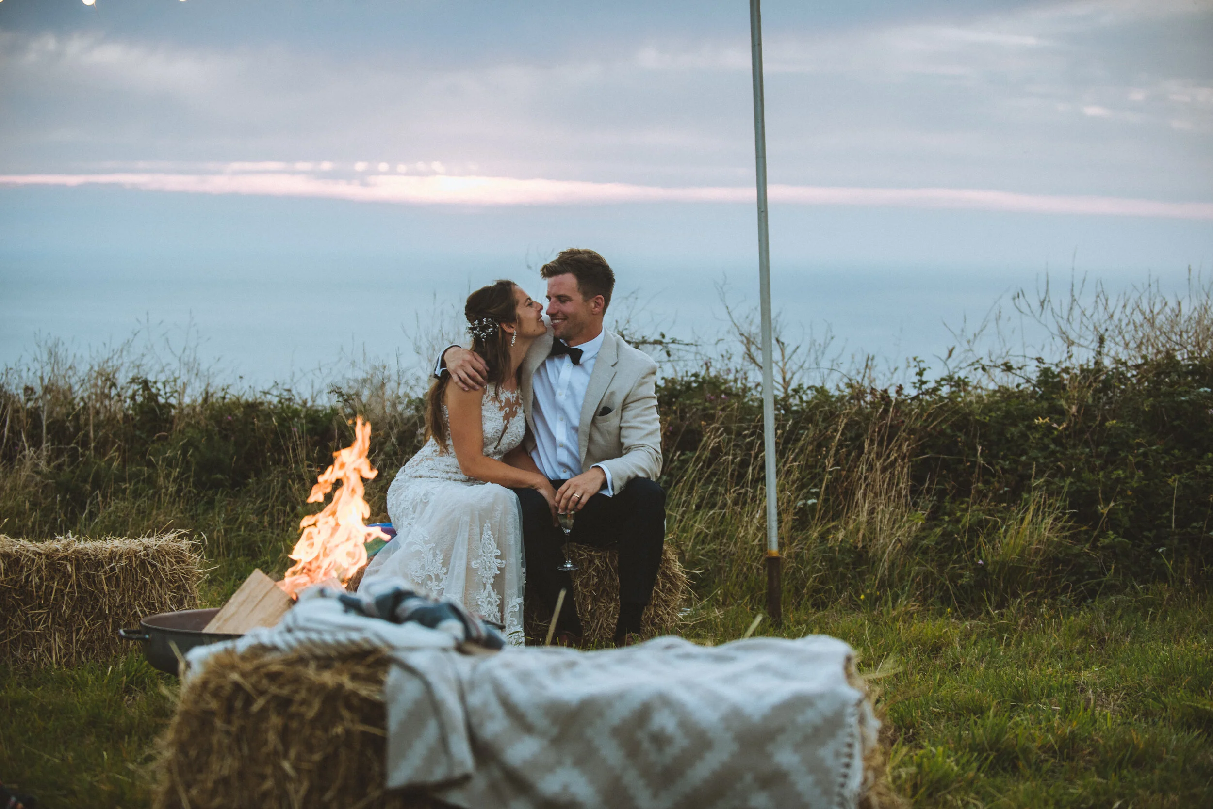 Daymer bay wedding photography in Cornwall captured by Mark Shaw Photography - A bride and groom sitting on hay bales outside during sunset, embracing and smiling, with a small campfire in front of them and a flagpole nearby.