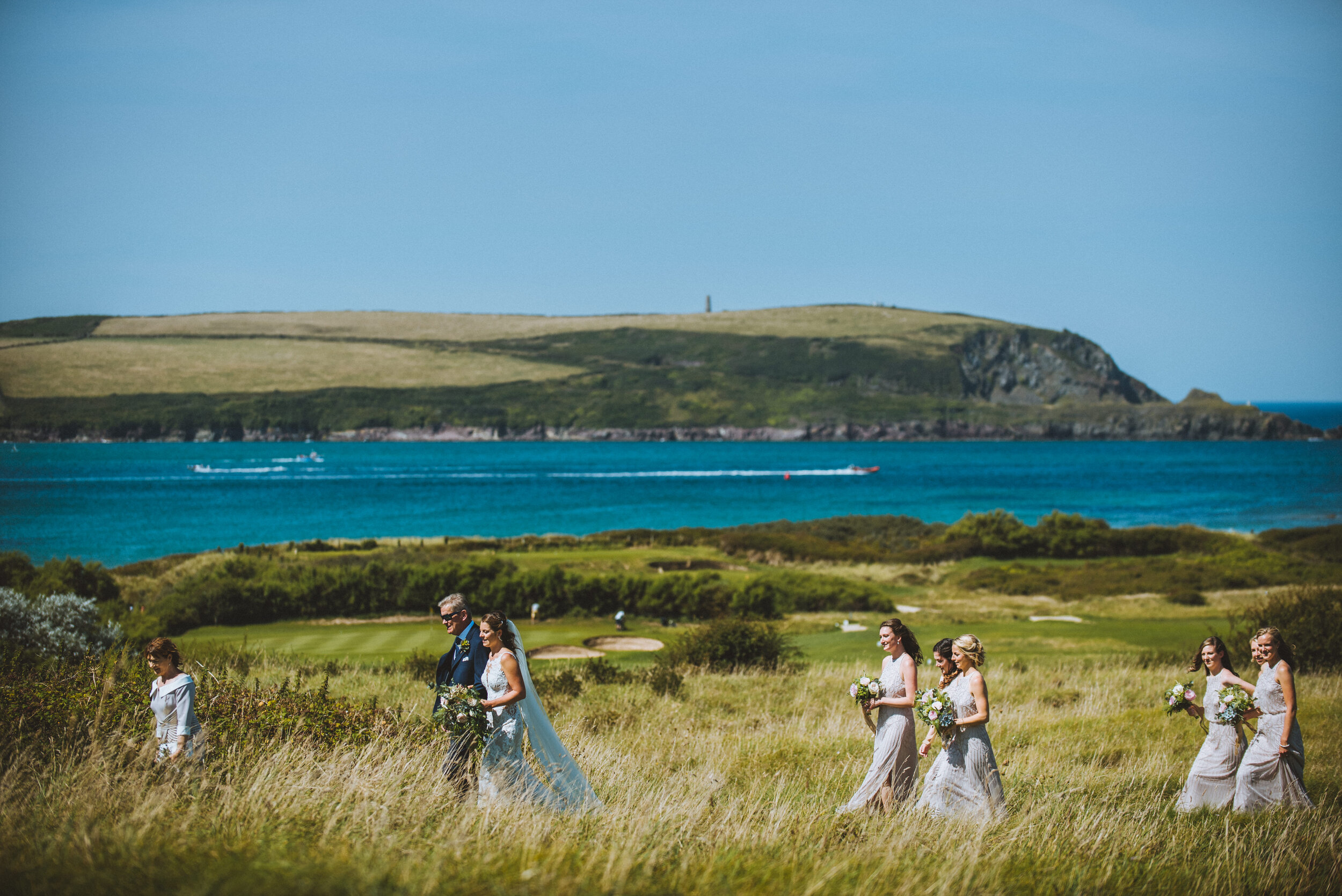 Daymer bay wedding photography in Cornwall captured by Mark Shaw Photography - A wedding ceremony taking place outdoors on a sunny day with a grassy field, a body of water, and hills in the background.