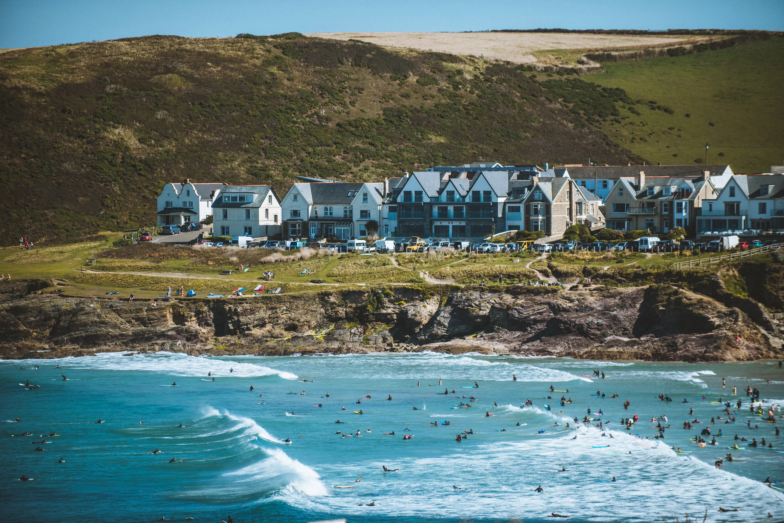 Beach with many surfers and people enjoying the sunny day, lined by rocky cliffs and houses on a hillside.