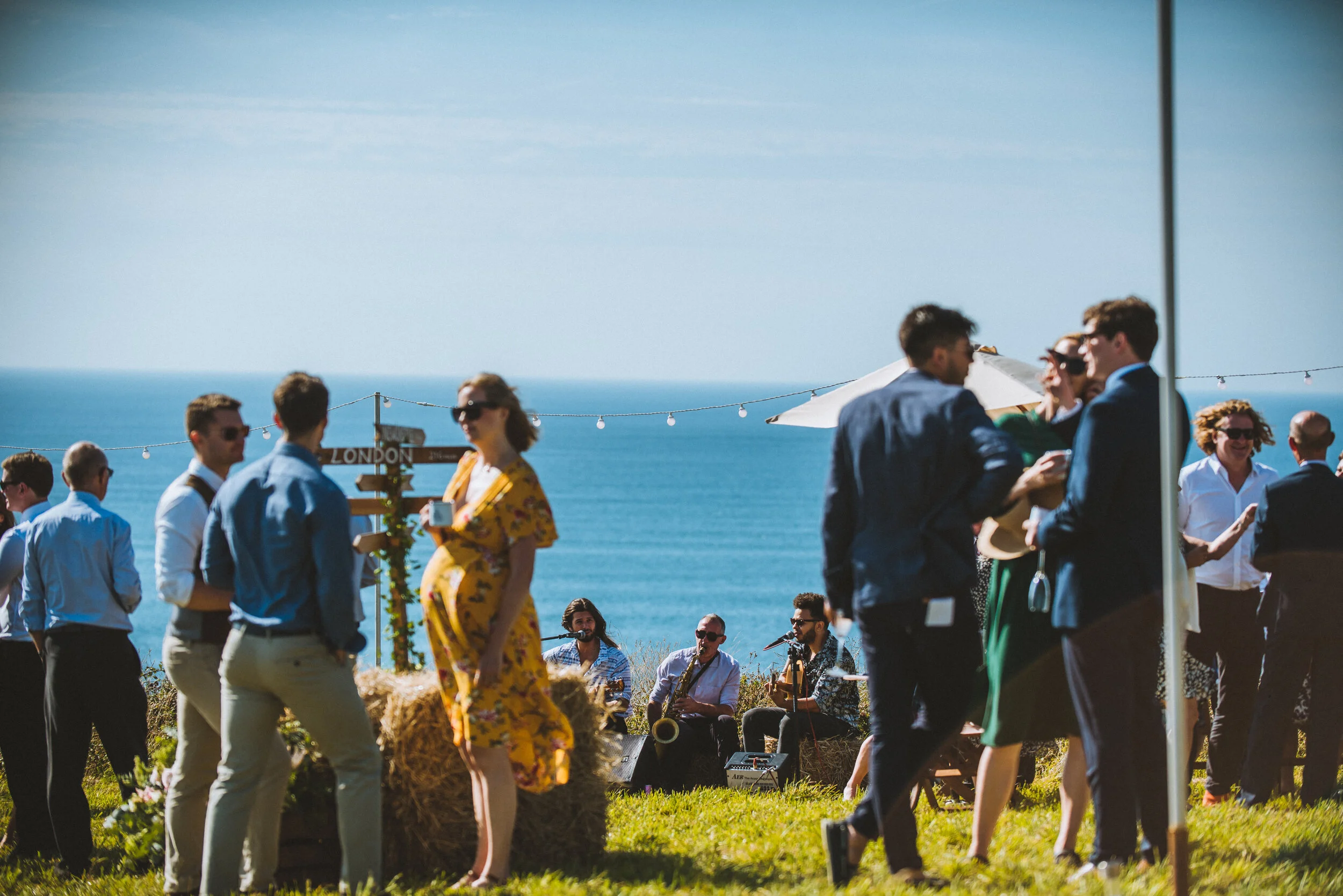 Wedding photo at Greenaway Field in Cornwall. People socializing and listening to live music outdoors near the ocean on a sunny day.
