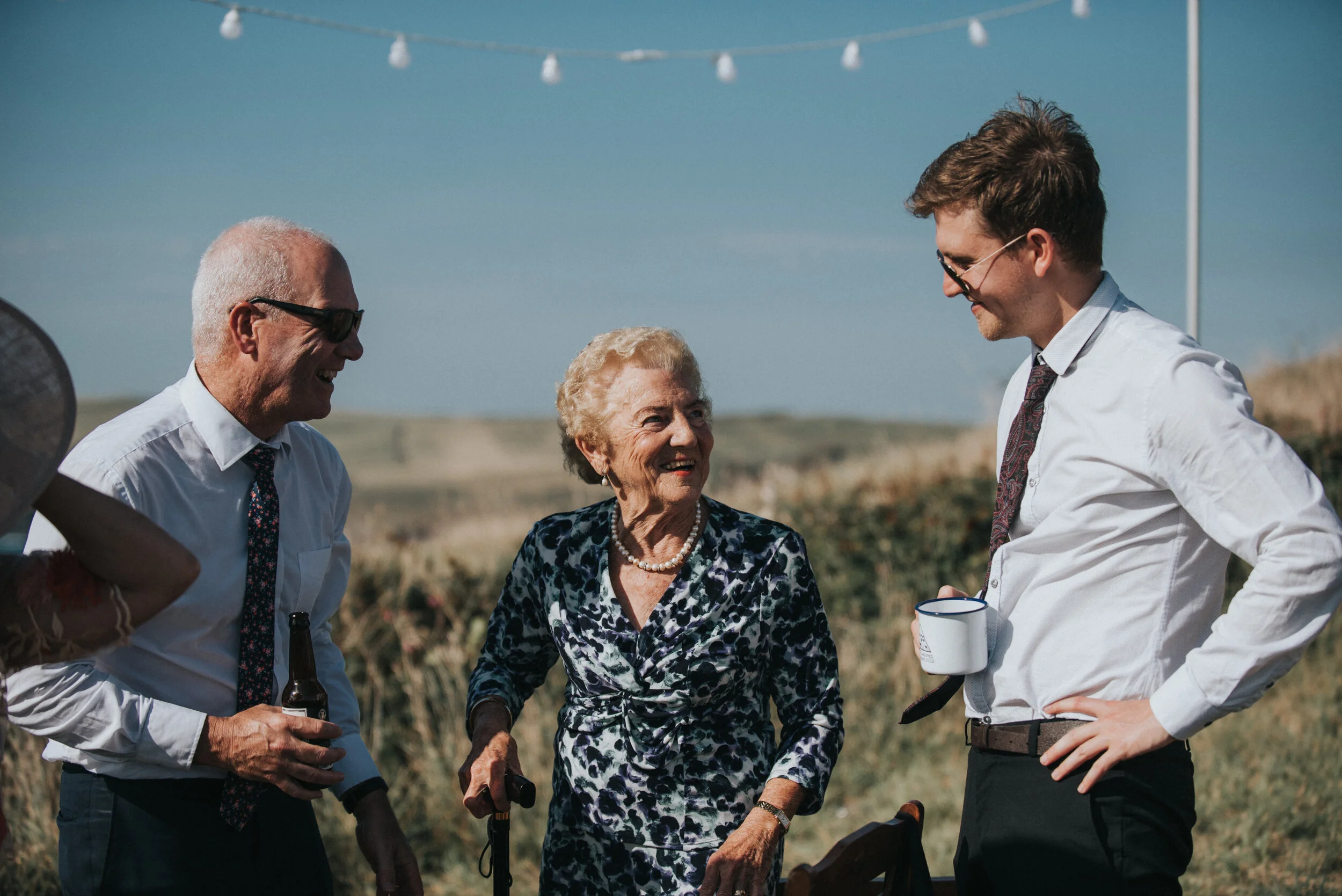 Four people talking outdoors on a sunny day. An elderly woman with a cane is in the center, smiling at a young man with glasses and a white shirt. An older man with sunglasses and a tie is also smiling, holding a beer bottle. In the background, there