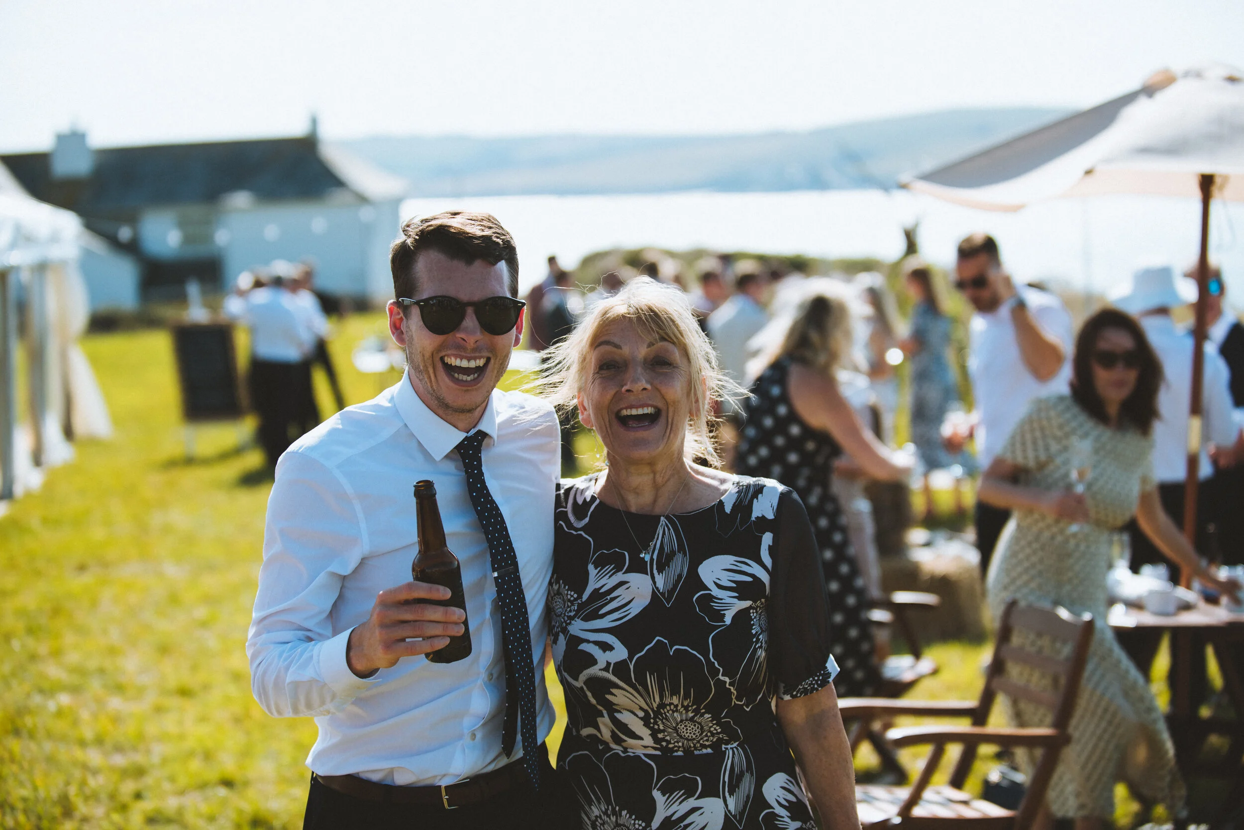Two people smiling, a young man wearing sunglasses and a woman, at an outdoor gathering near water with others in the background.