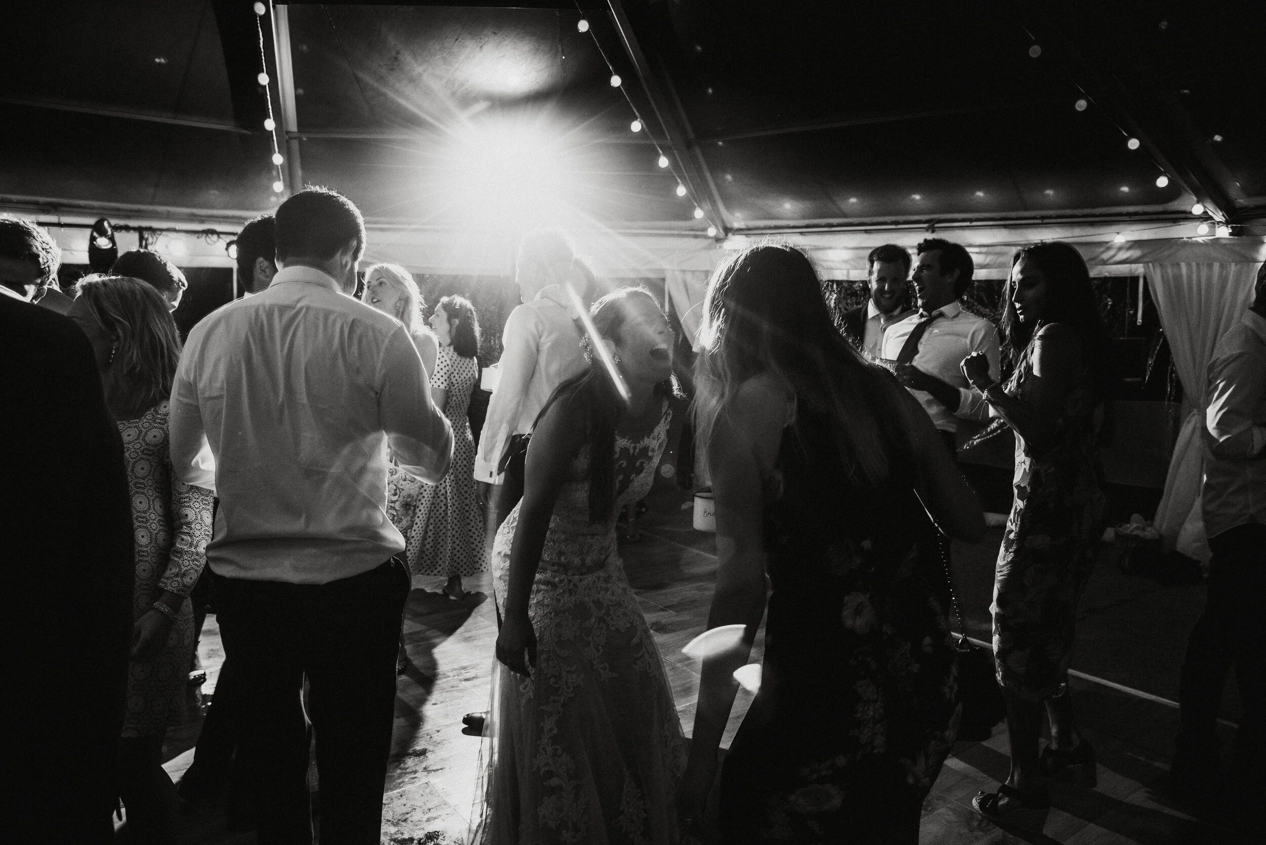 Daymer bay wedding photography captured by Cornwall wedding photographer Mark Shaw Photography -People dancing and socializing at a party or celebration under a tent with string lights, black and white photo.