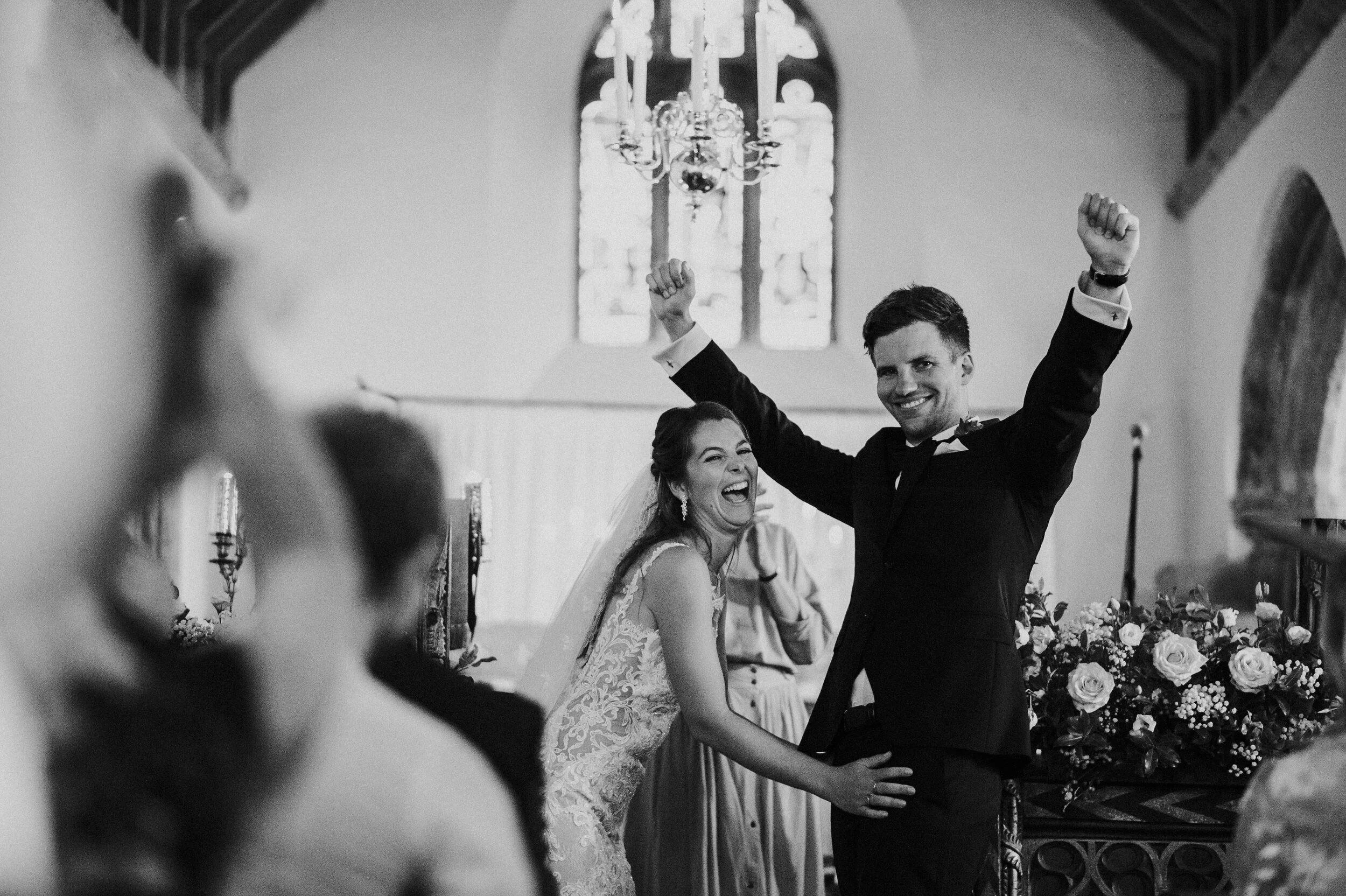 St Enodoc Church wedding photograph, Daymer Bay in A bride and groom celebrating during a wedding ceremony inside a church, raising their arms in joy, with the bride laughing and the groom smiling, surrounded by wedding guests and floral decorations.