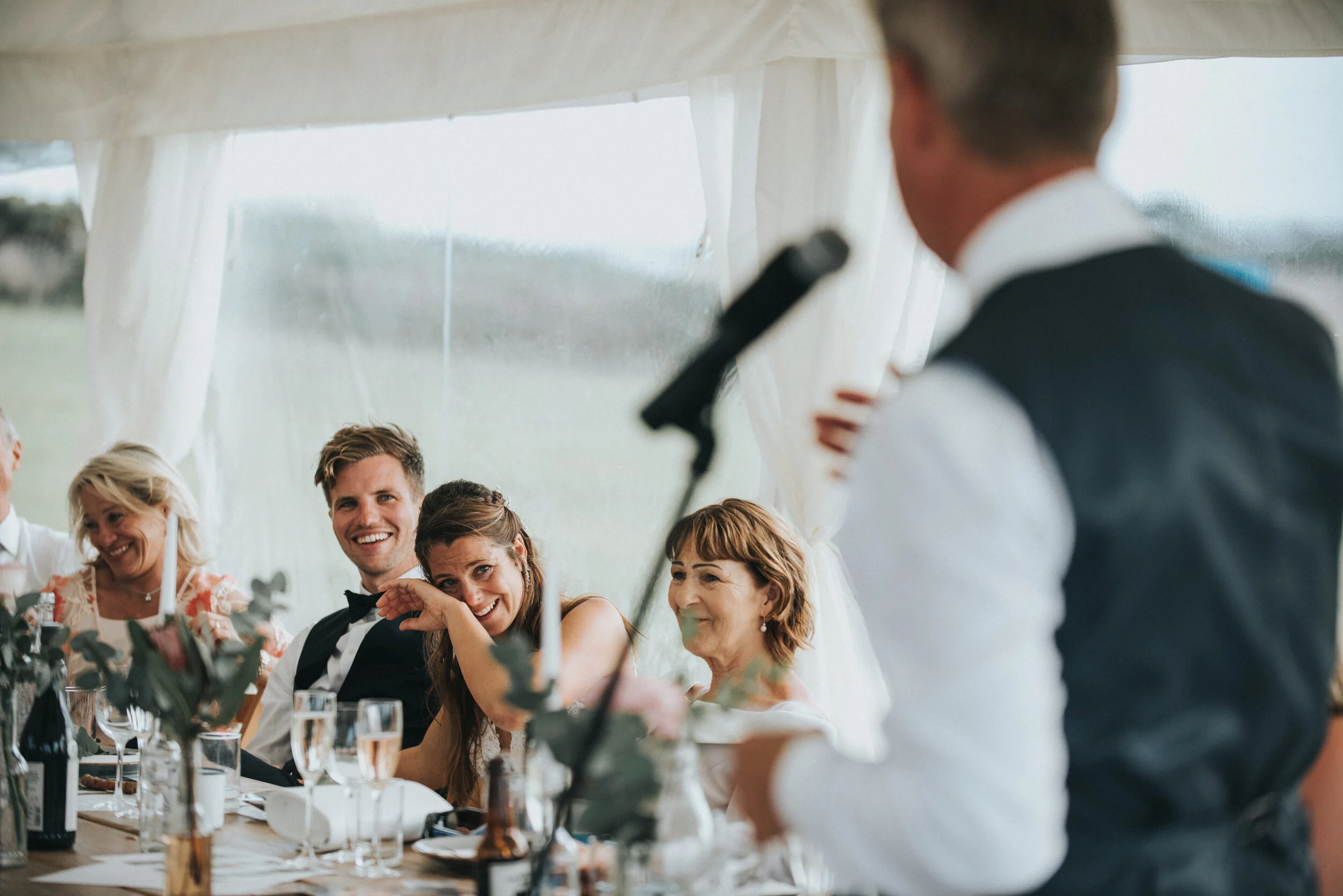 Daymer bay wedding photography in Cornwall captured by Mark Shaw Photography - Guests at a wedding reception sit at a table inside a tent, smiling and listening to a man giving a speech or toast.