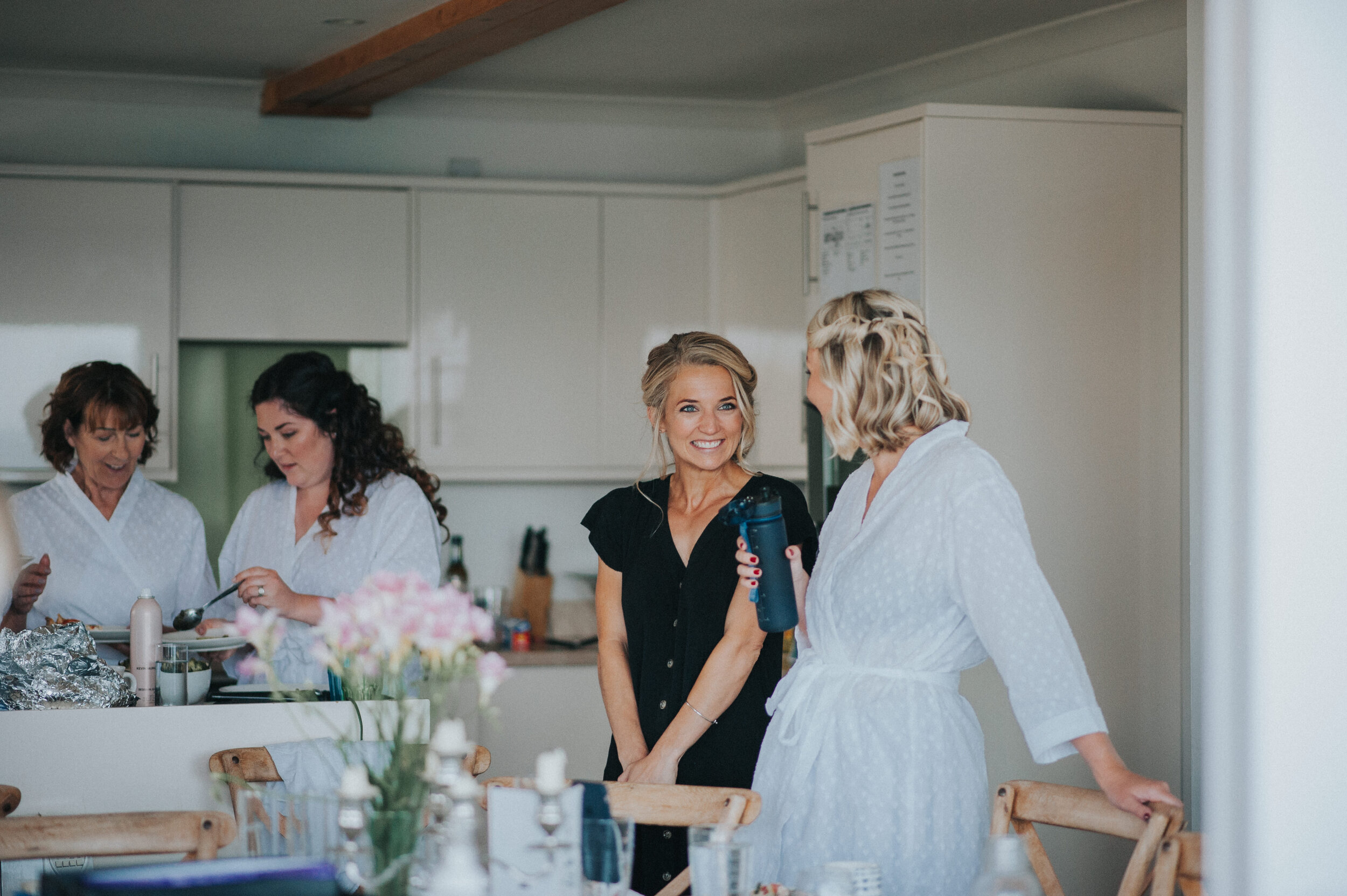 Women gather in a kitchen, chatting and preparing food, with one woman holding a water bottle.
