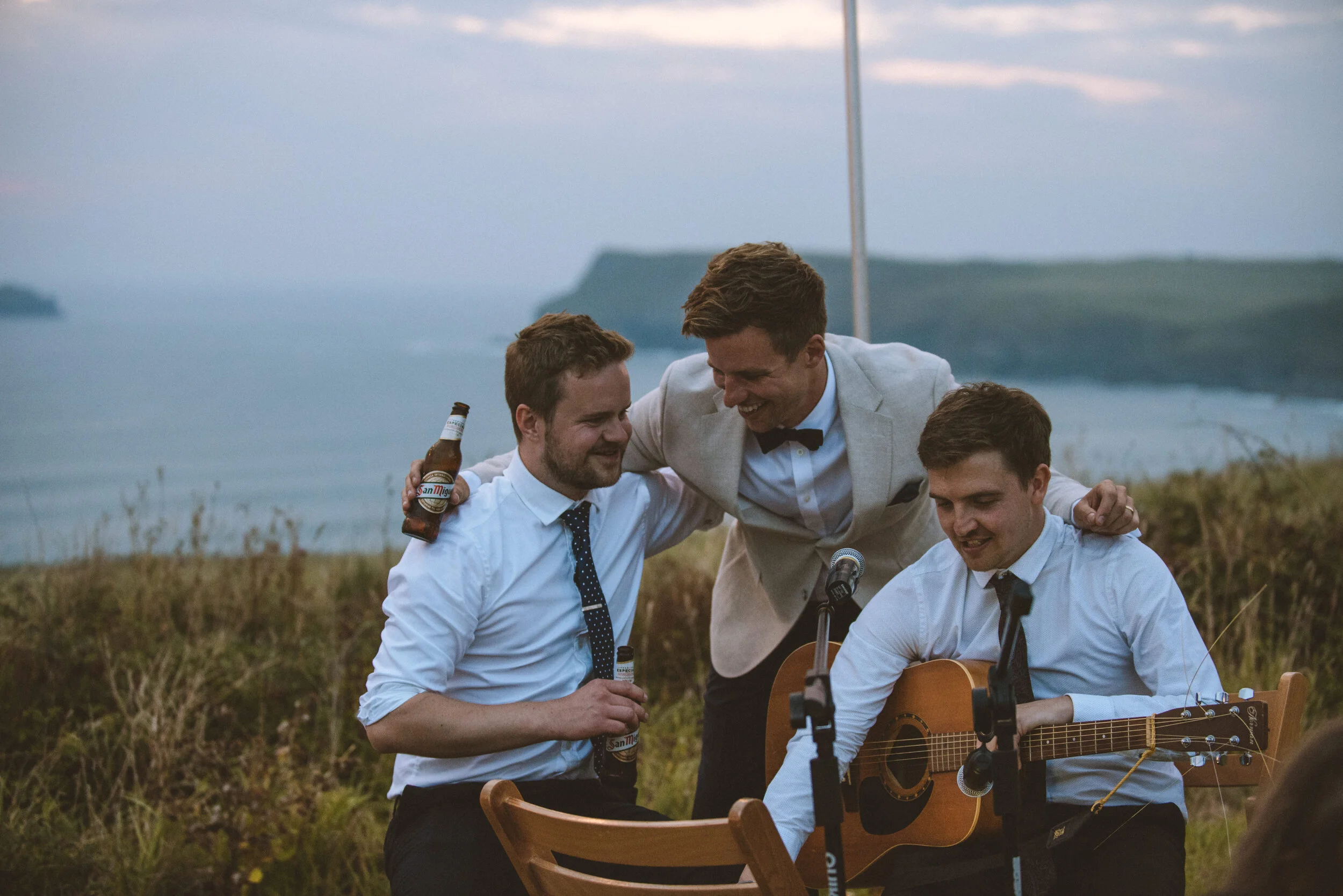 Daymer bay wedding photography in Cornwall captured by Mark Shaw PhotogThree young men dressed formally, with two holding beer bottles and one playing guitar, are gathered outdoors near the coast at sunset, smiling and enjoying a musical performance.