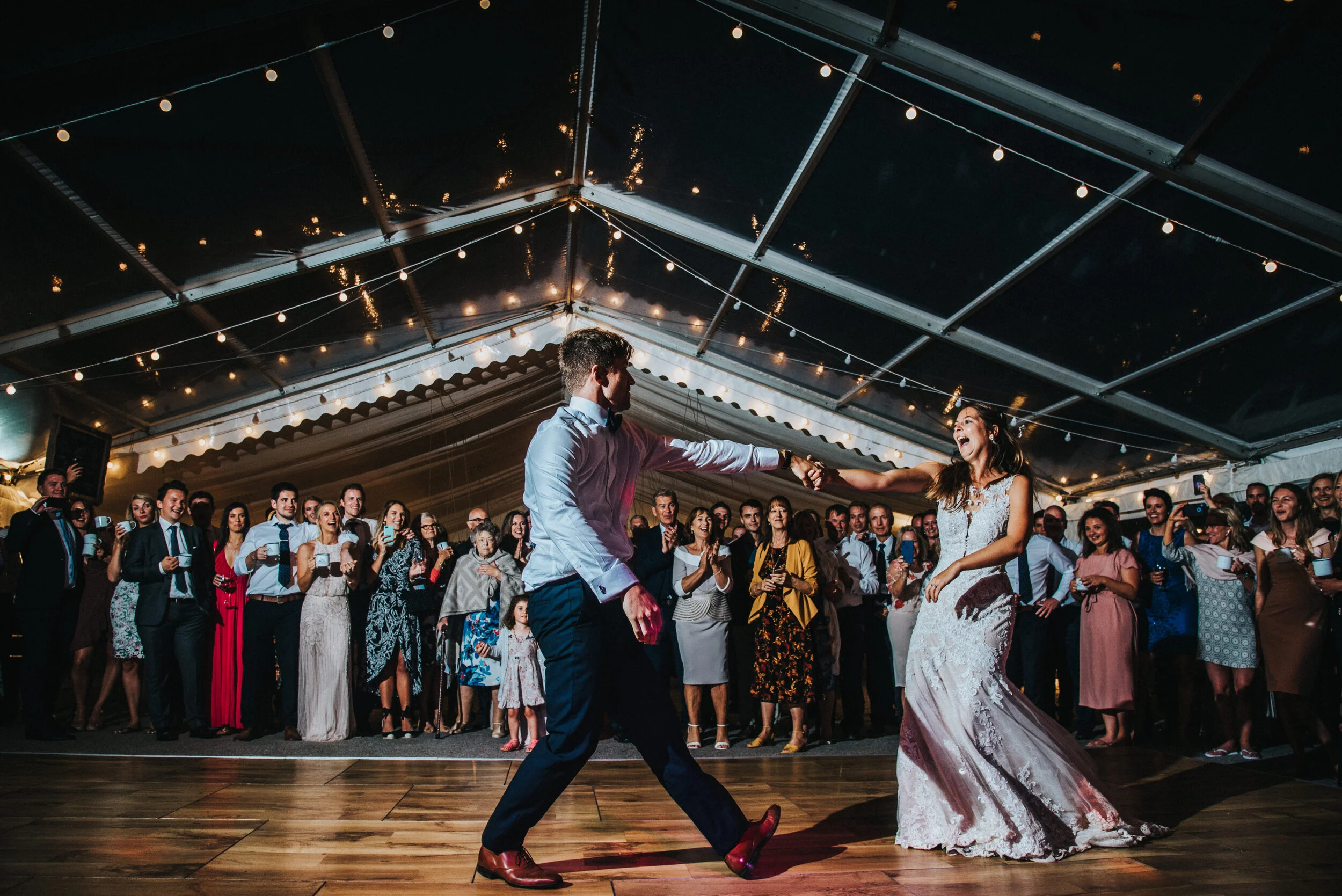 Daymer bay wedding photography captured by Cornwall wedding photographer Mark Shaw Photography - first dance photo. A bride and groom dancing at their wedding reception under a tent with string lights, surrounded by guests watching and cheering.