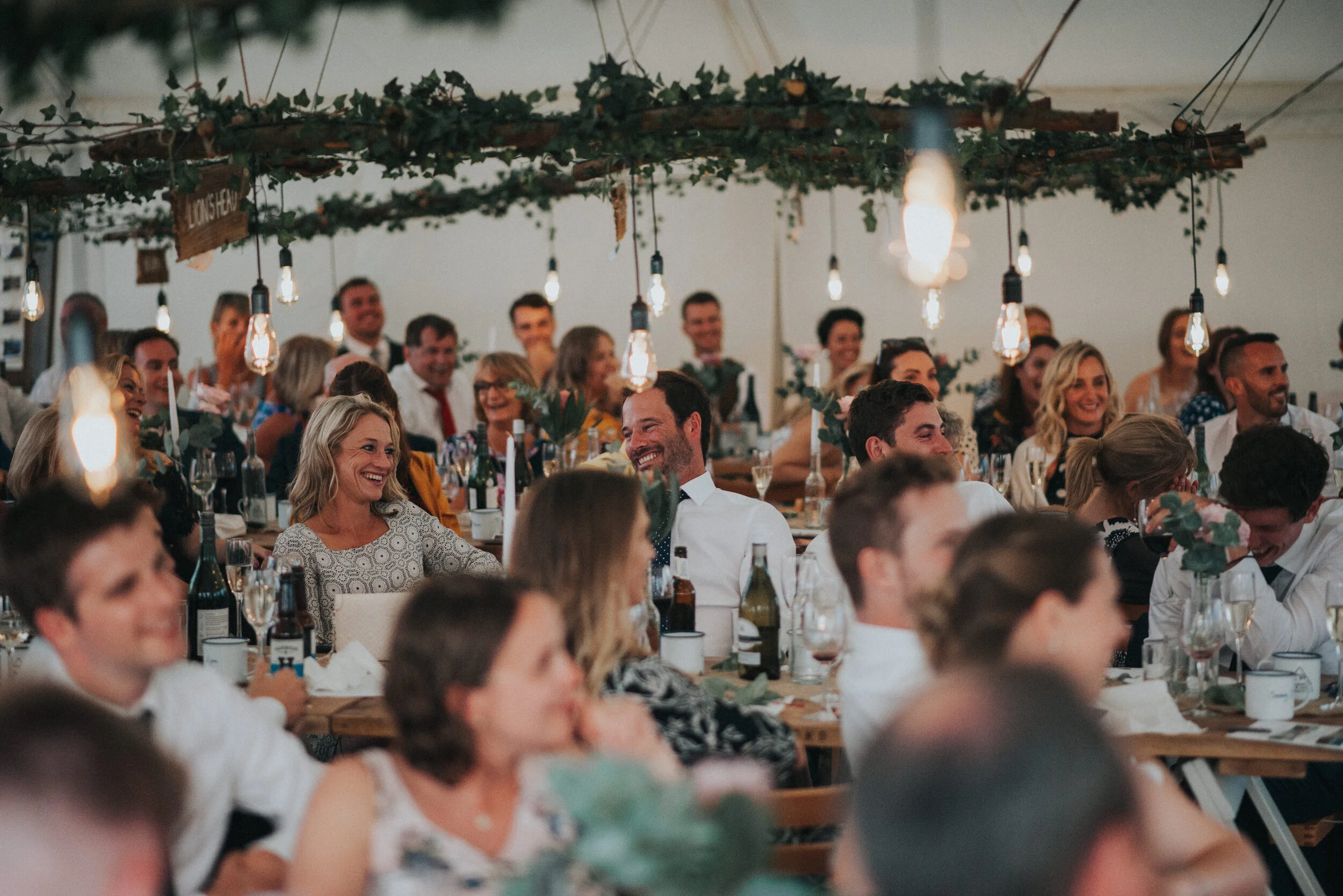Daymer bay wedding photography in Cornwall captured by Mark Shaw Photography - Guests at a wedding reception seated at decorated tables, laughing and chatting under hanging lights and greenery.