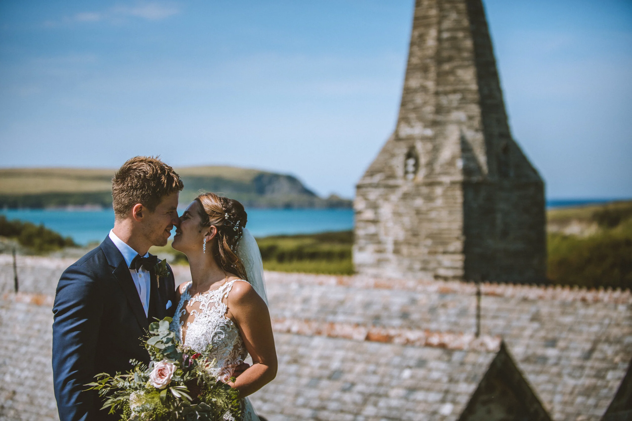 St Enodoc Church wedding photograph, Daymer Bay in Cornwall. A bride and groom standing close together outdoors with a stone tower and body of water in the background.