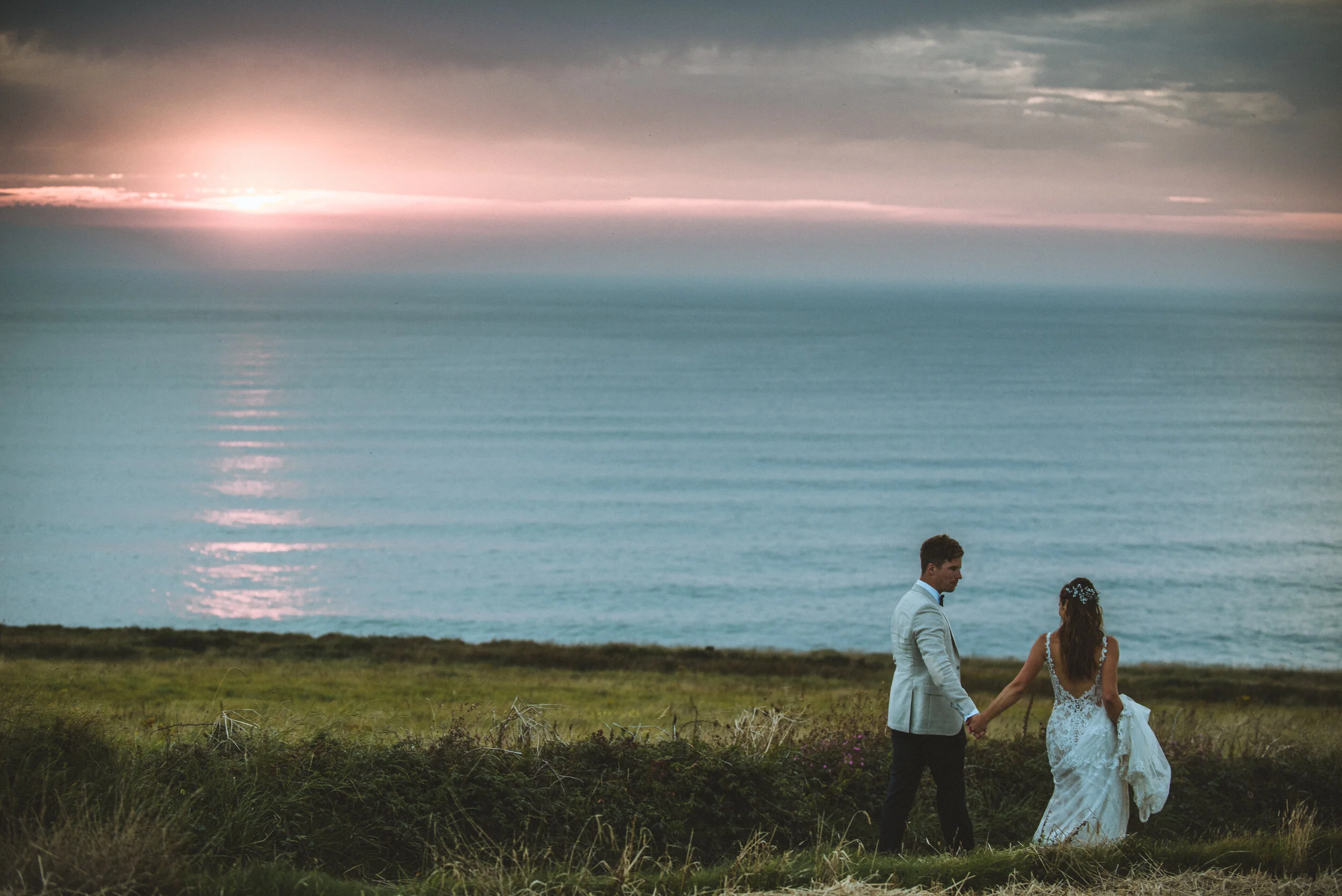 Daymer bay wedding photography in Cornwall captured by Mark Shaw Photography - A bride and groom holding hands and walking toward the ocean at sunset, with a cloudy sky and calm water in the background.