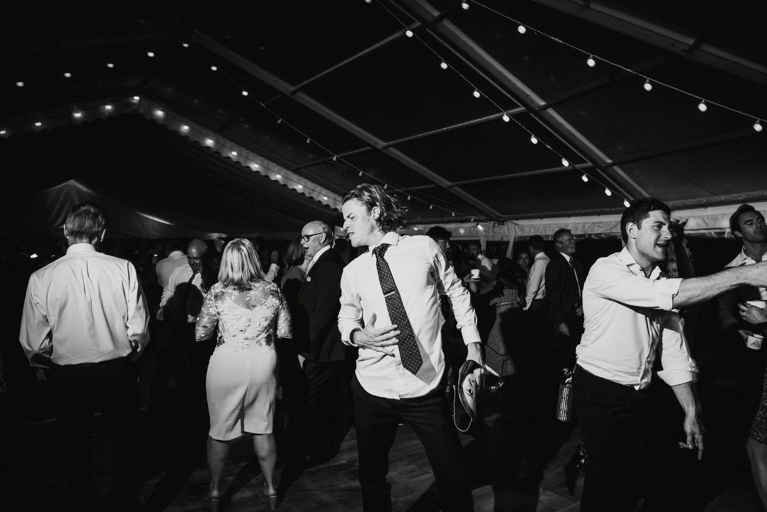 Daymer bay wedding photography captured by Cornwall wedding photographer Mark Shaw Photography -People dancing and socializing at an indoor party or reception, with string lights overhead, in black and white.