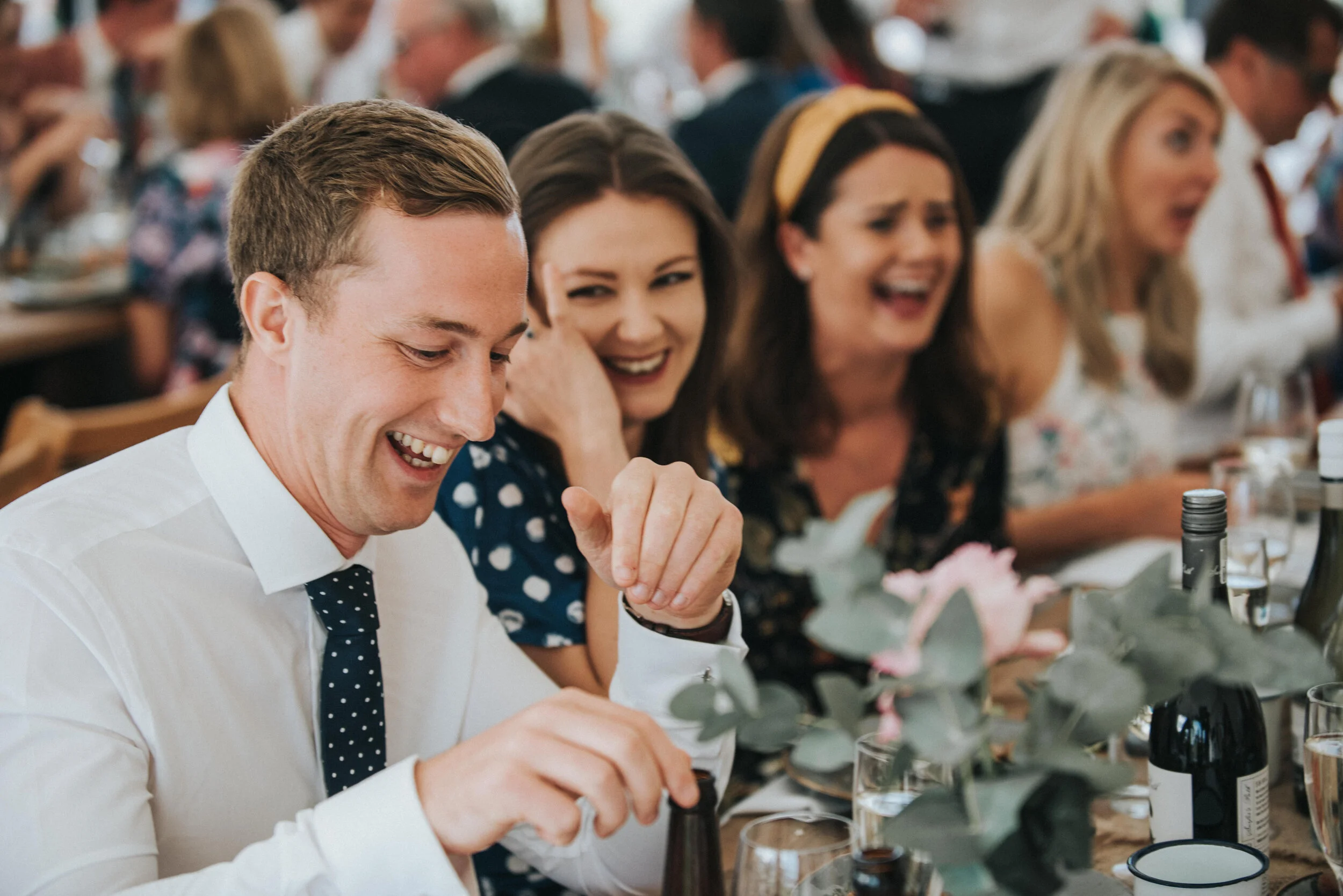 Daymer bay wedding photography in Cornwall captured by Mark Shaw Photography - People laughing and enjoying a meal at a social gathering or party, sitting at a decorated table with flowers and drinks.