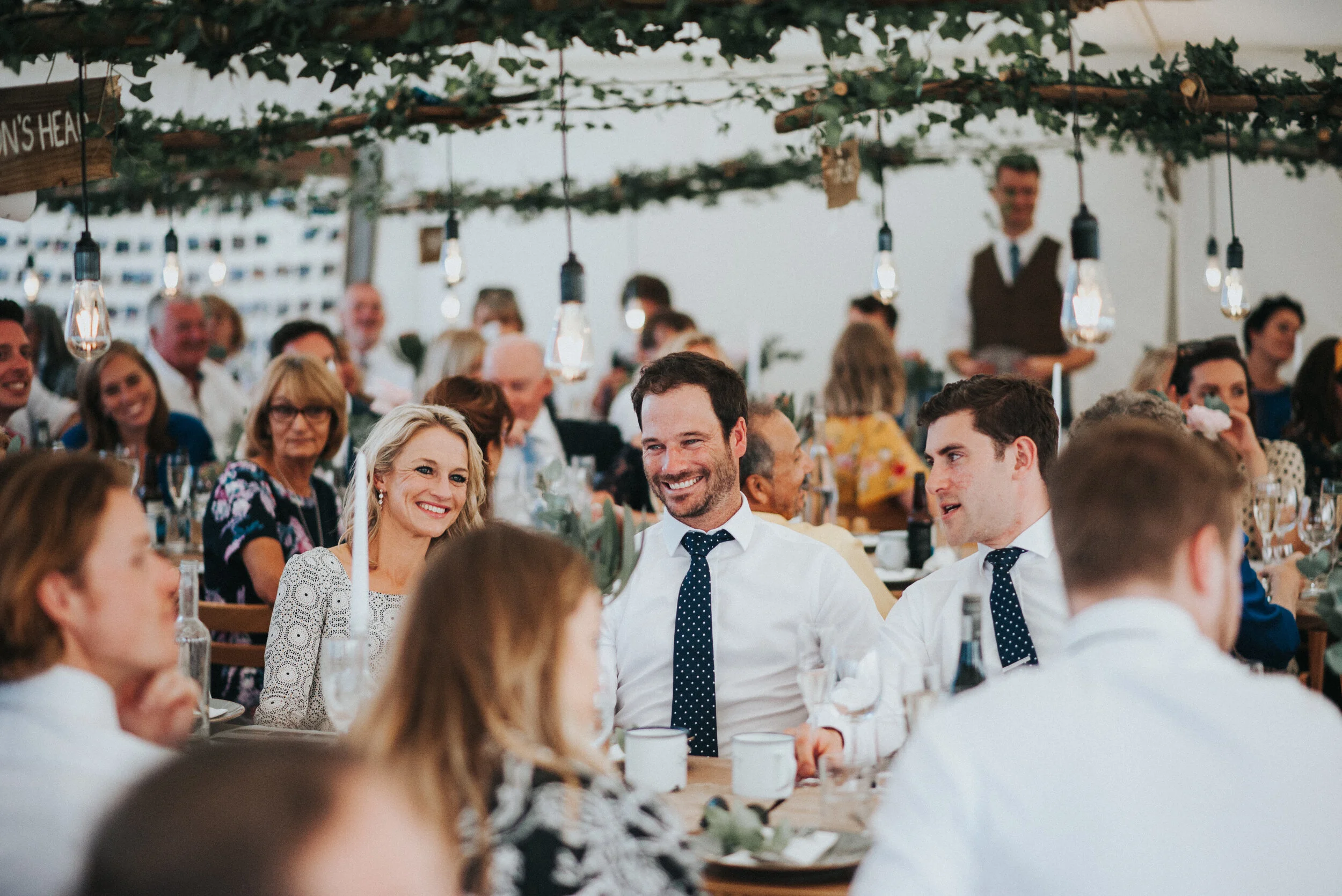 Daymer bay wedding photography in Cornwall captured by Mark Shaw Photography - Guests at a wedding reception seated at tables, smiling and enjoying the event under string lights and greenery decor.