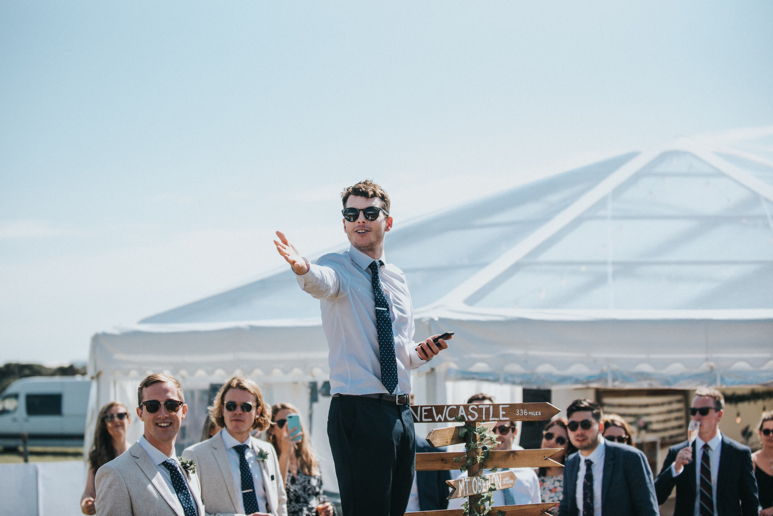 Wedding photo at Greenaway Field in Cornwall. Man in sunglasses delivering a speech at an outdoor wedding, with guests in formal attire watching, and a sign pointing to Newcastle in the background.