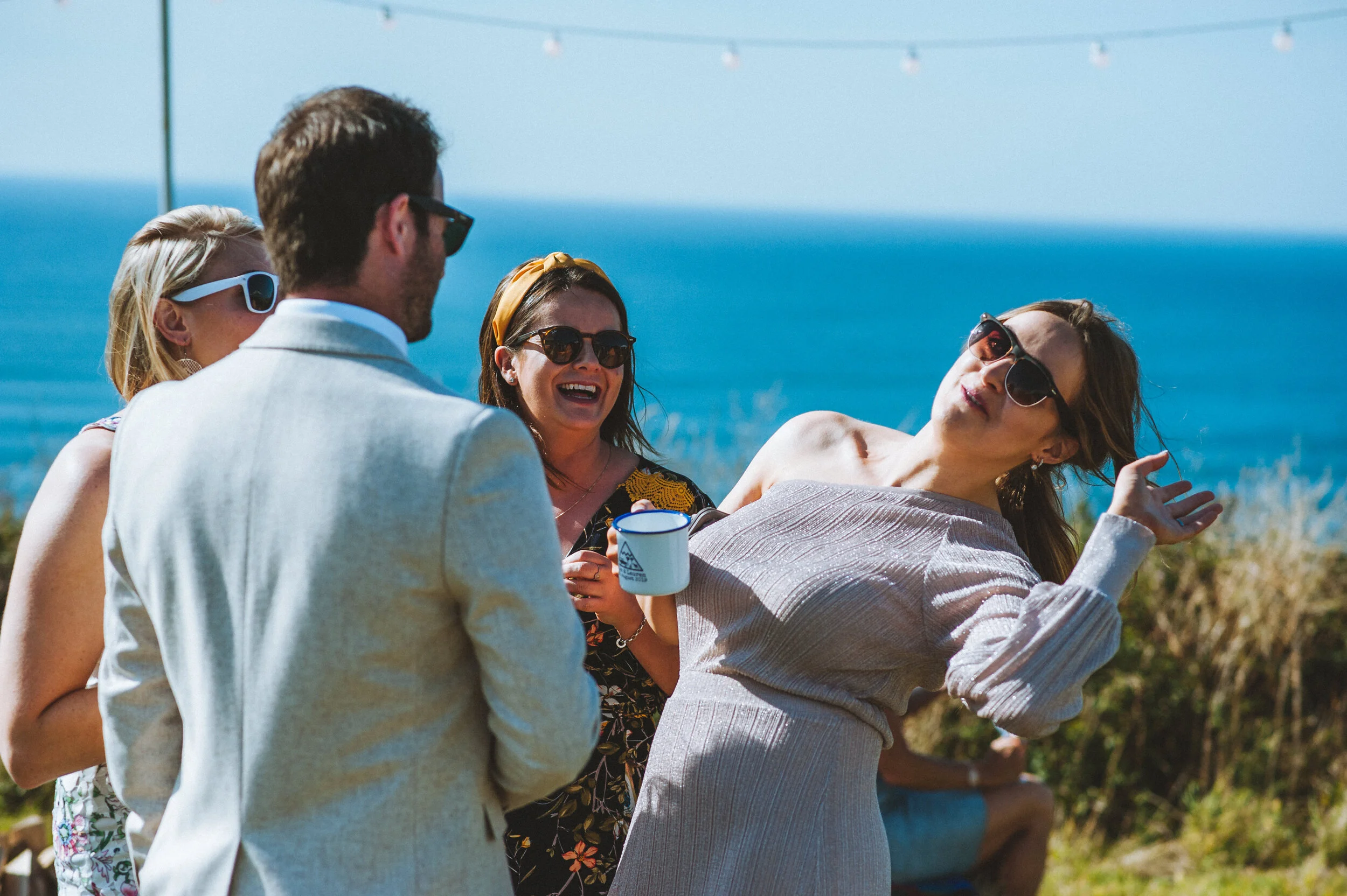 Wedding photo at Greenaway Field in Cornwall. Group of people enjoying outdoors near the ocean, laughing and wearing sunglasses, with some holding mugs.