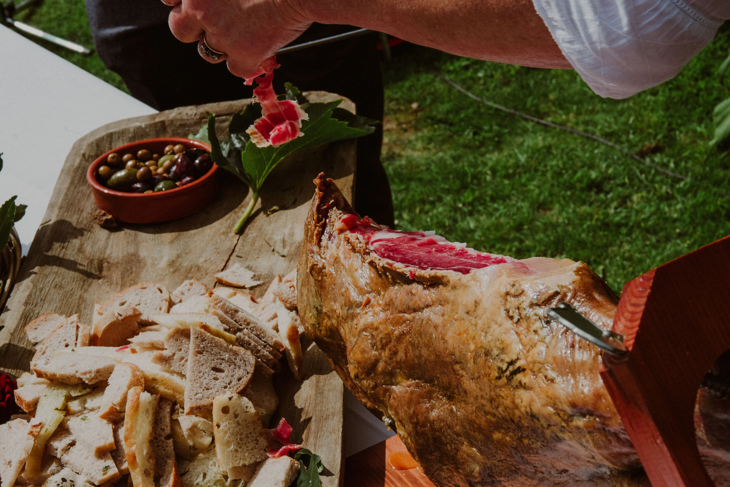 Person slicing cured ham with plates of bread and olives on a wooden board outdoors.