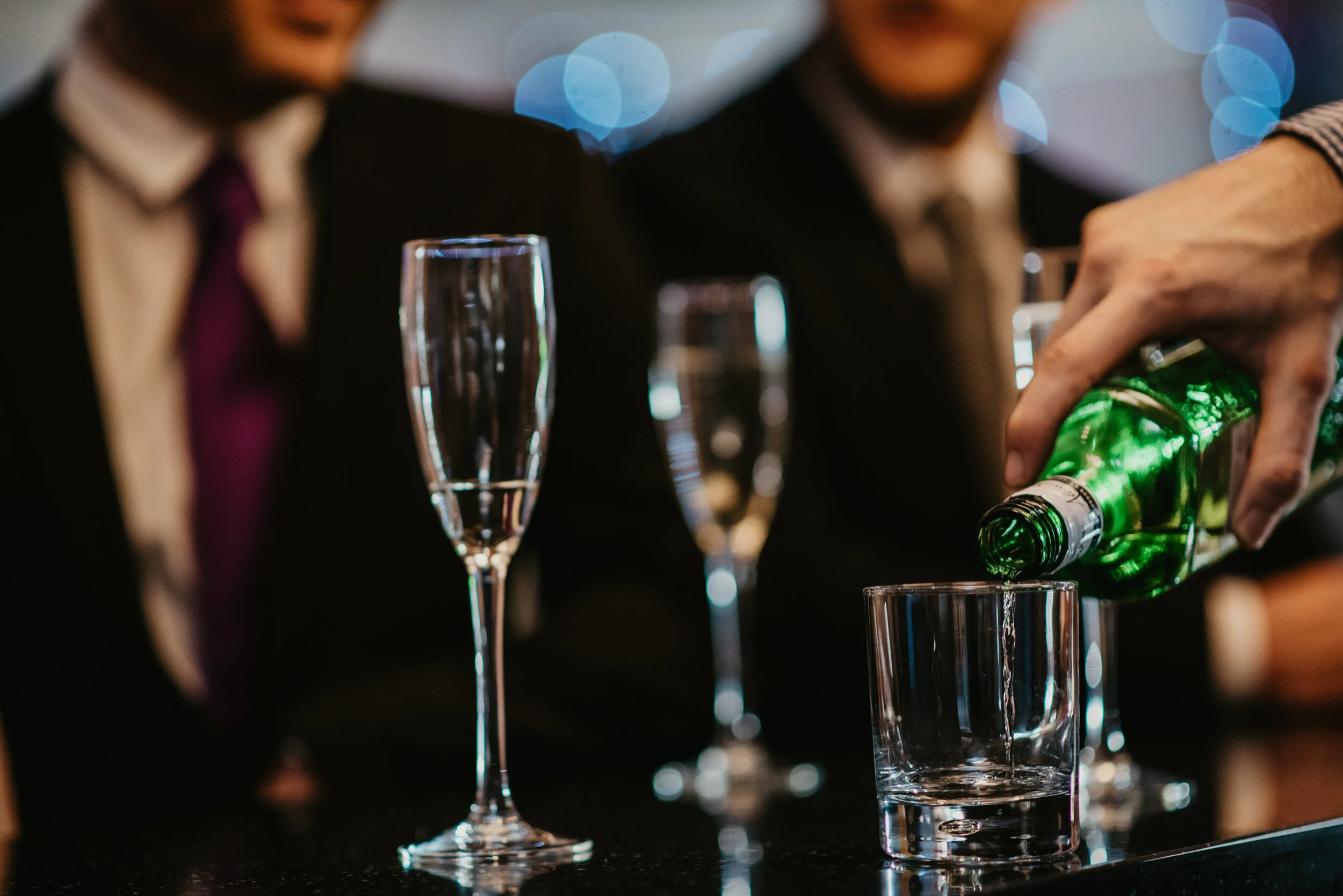 Bartender pouring liquid into glass, champagne flutes in background, people in suits.