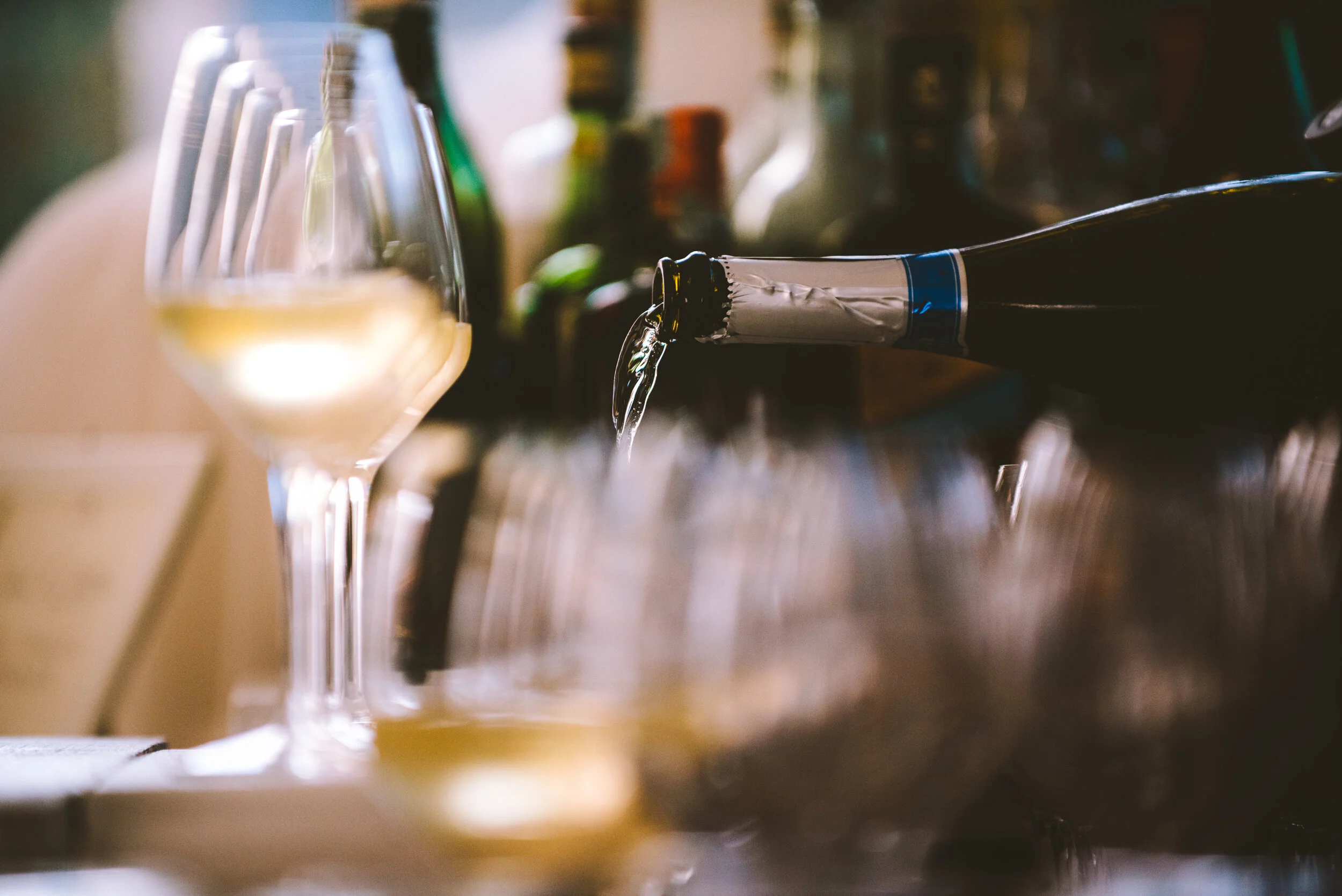 Wine being poured into a glass with multiple wine glasses and bottles in the background.