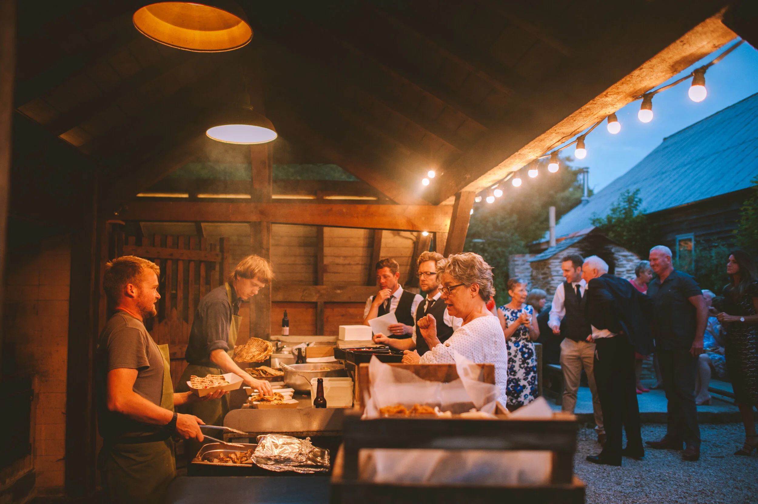 Outdoor evening gathering with people near a food counter, warm lighting, event setting.