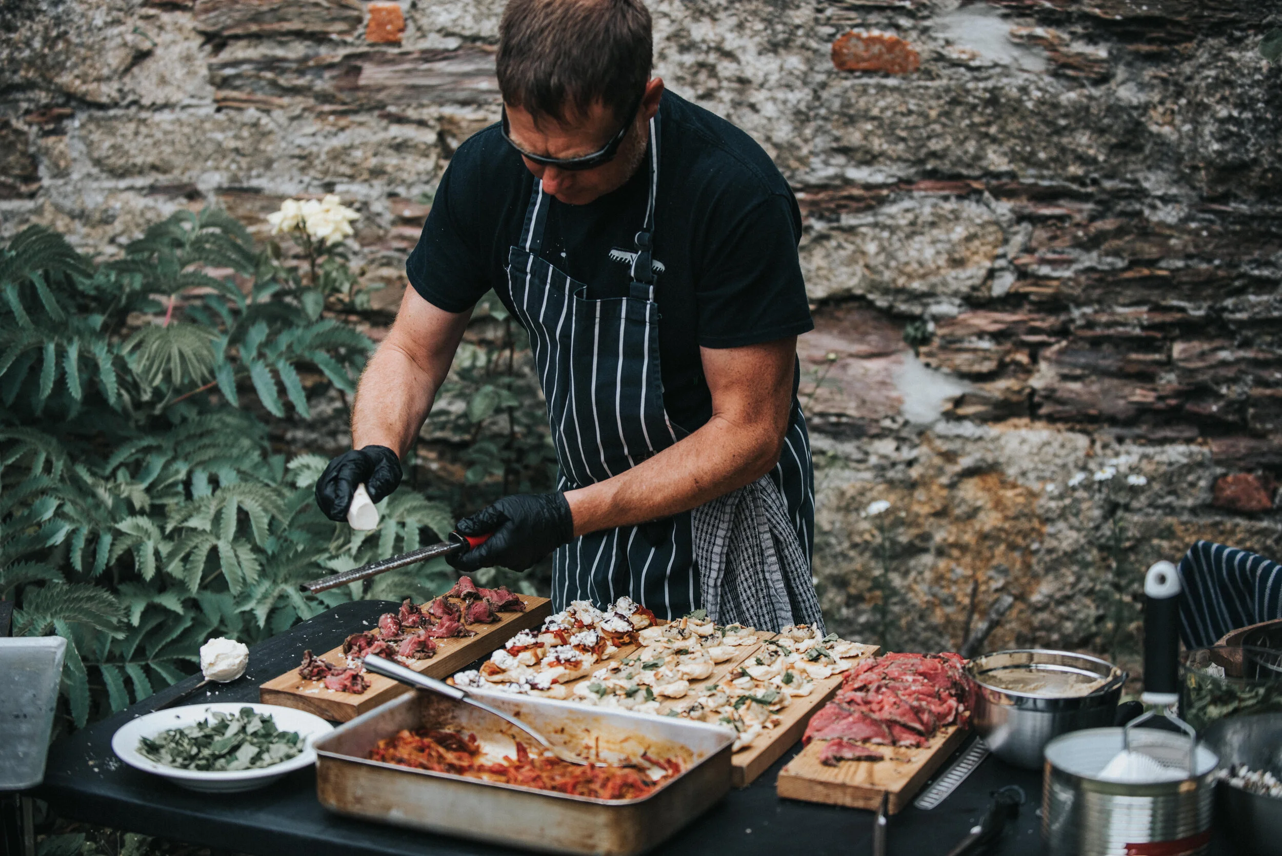 Chef in apron preparing food outdoors on a table with various ingredients and dishes.