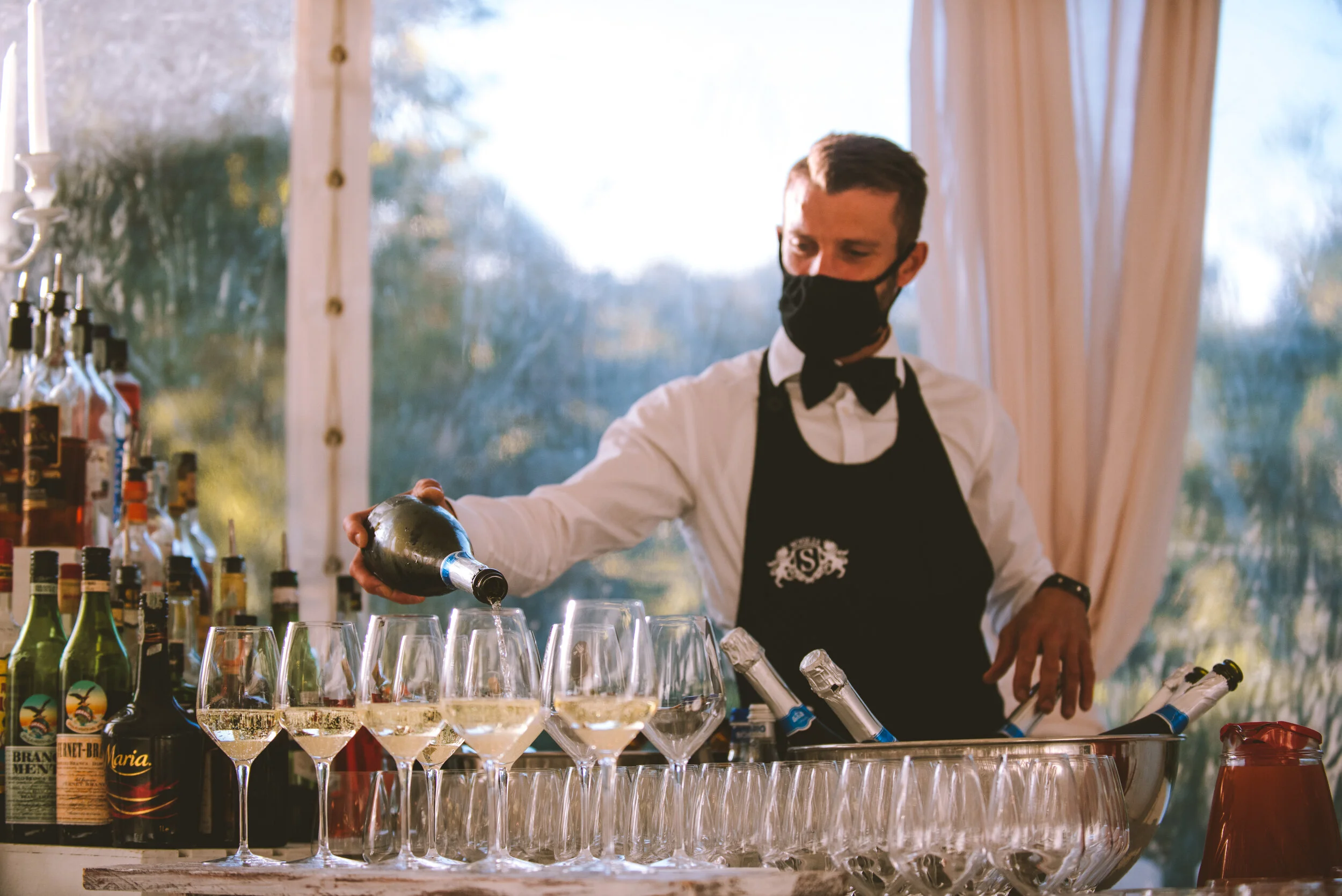 Bartender wearing a black mask and apron pouring white wine into glasses at a bar counter, with various bottles in the background.