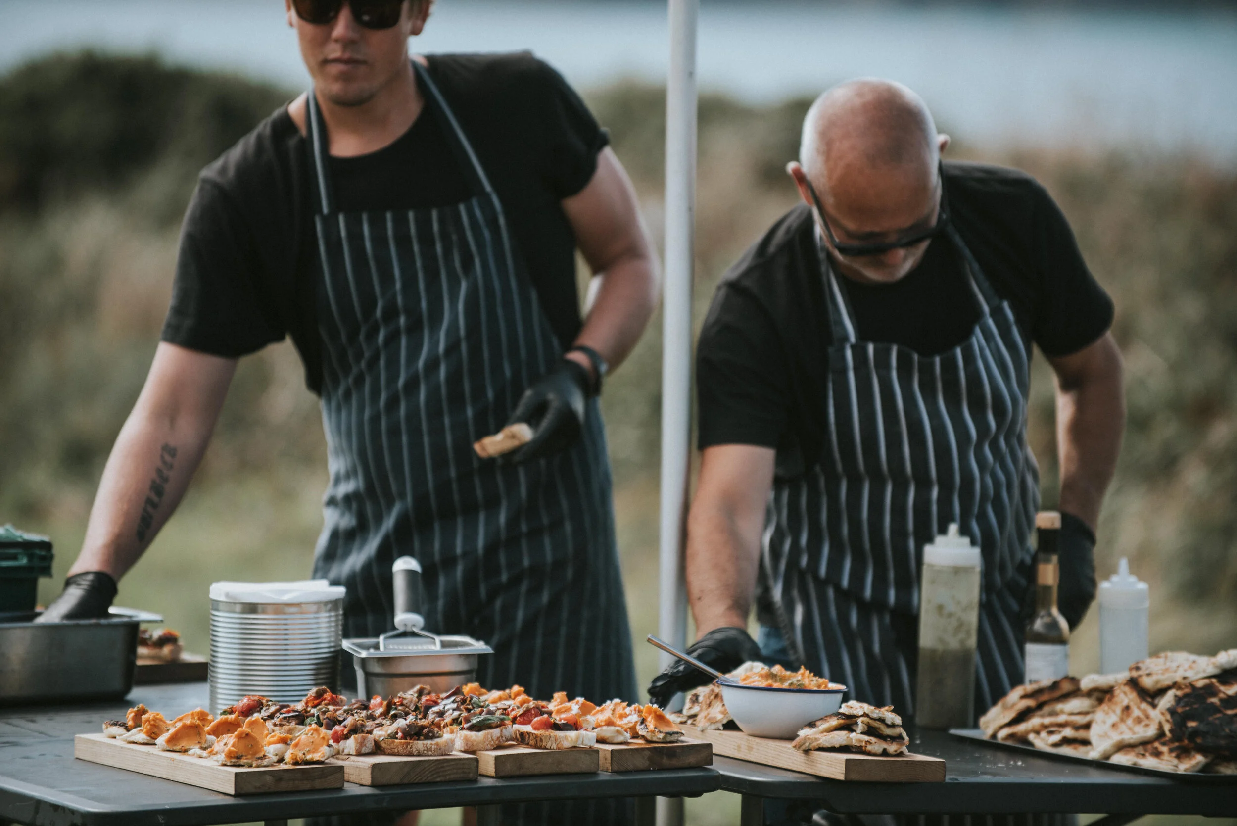 Two chefs in striped aprons preparing food on a table outdoors, with trays of appetizers and condiments.