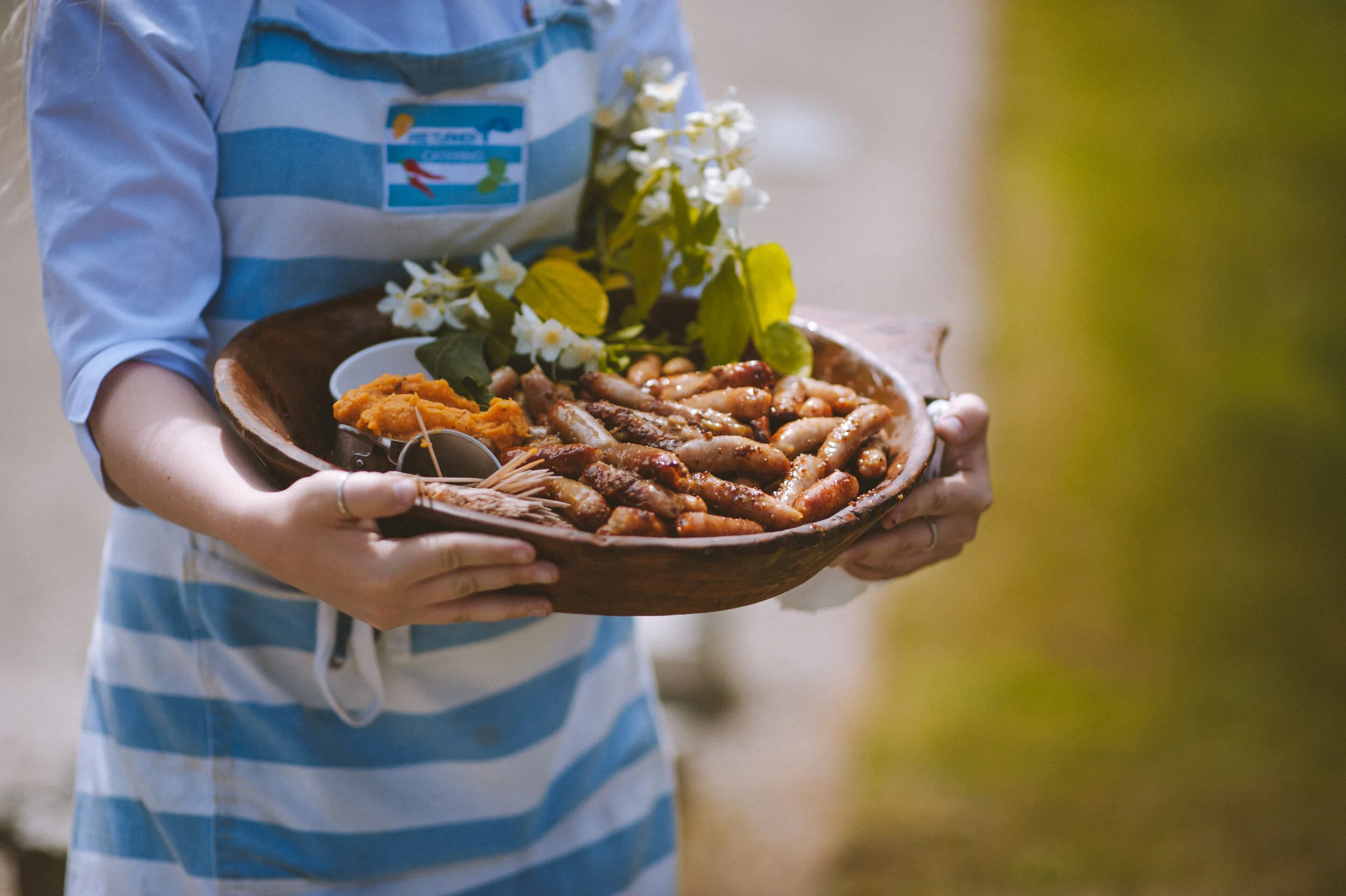Person holding a wooden bowl with small sausages, mashed vegetables, and flowers, wearing a striped apron.
