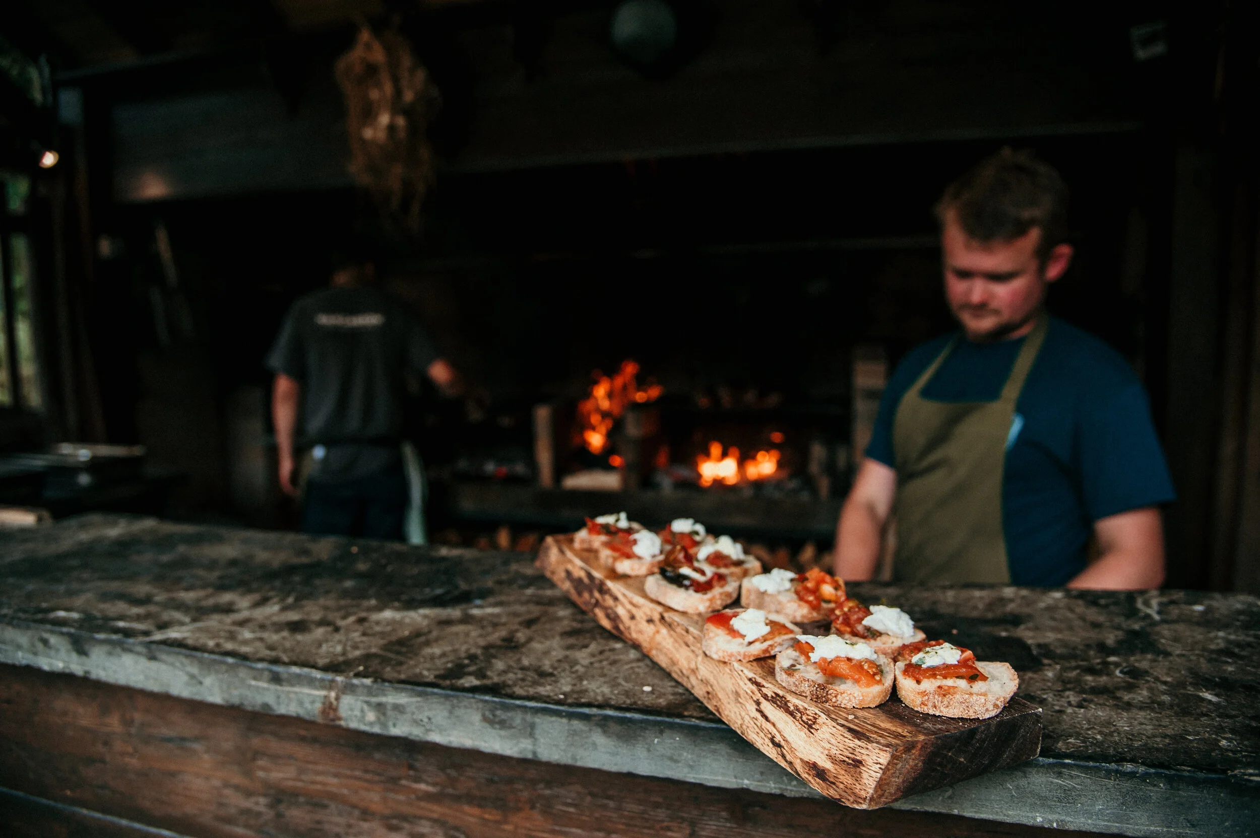 Outdoor kitchen with open fire in background, bread with toppings on wooden board in foreground, two chefs working, rustic setting.