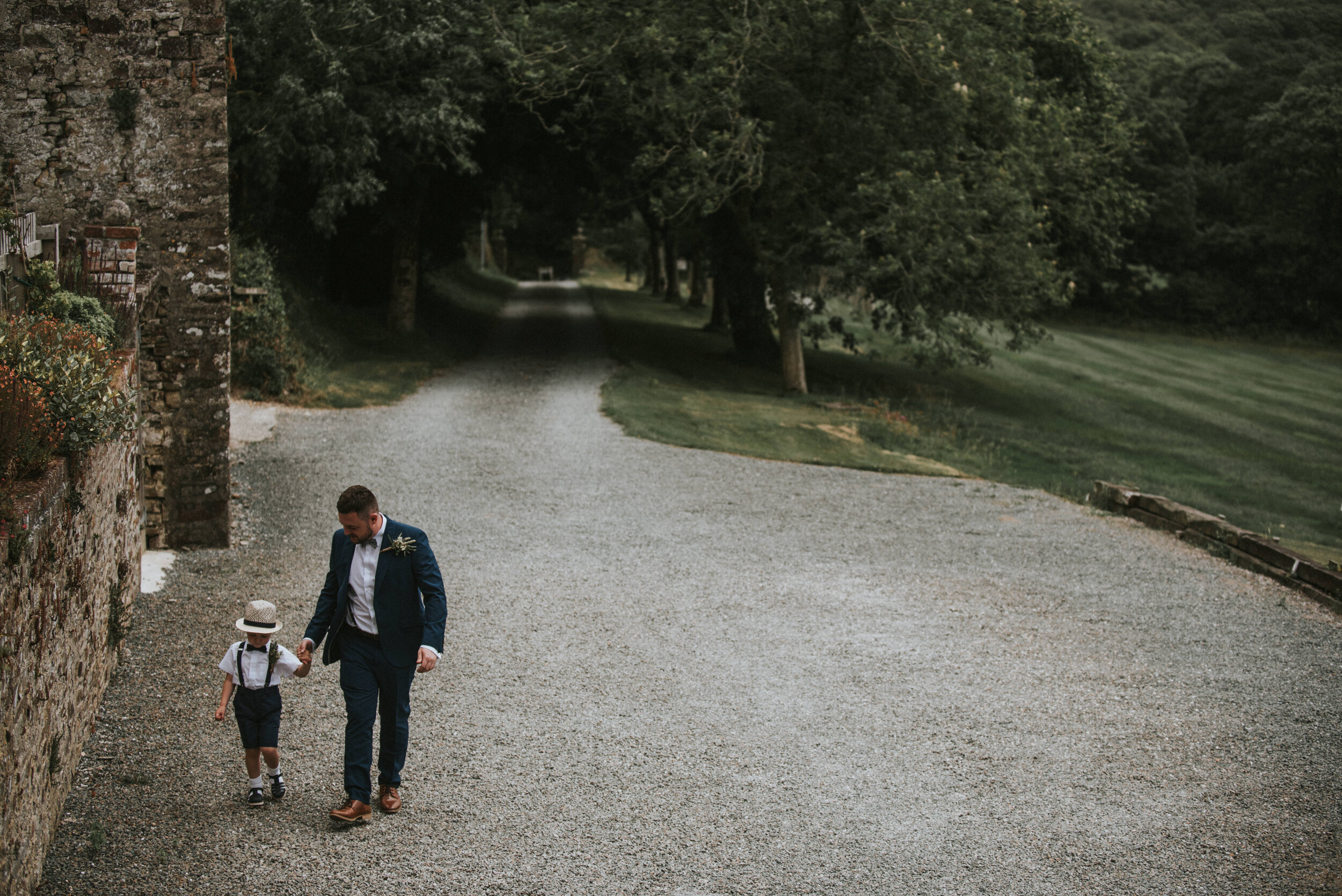 A man in a blue suit and a young boy in a hat and suspenders walk down a gravely pathway holding hands, with a tree-lined area and a stone wall to the left - Launcells Barton wedding photographer Cornwall