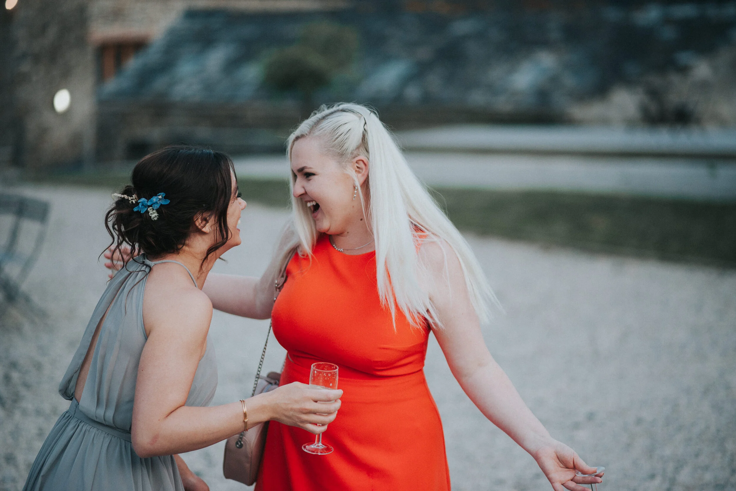 Two women smiling and laughing at an outdoor event, one in a gray dress with a blue hair accessory, and the other in a red dress holding a glass of champagne.