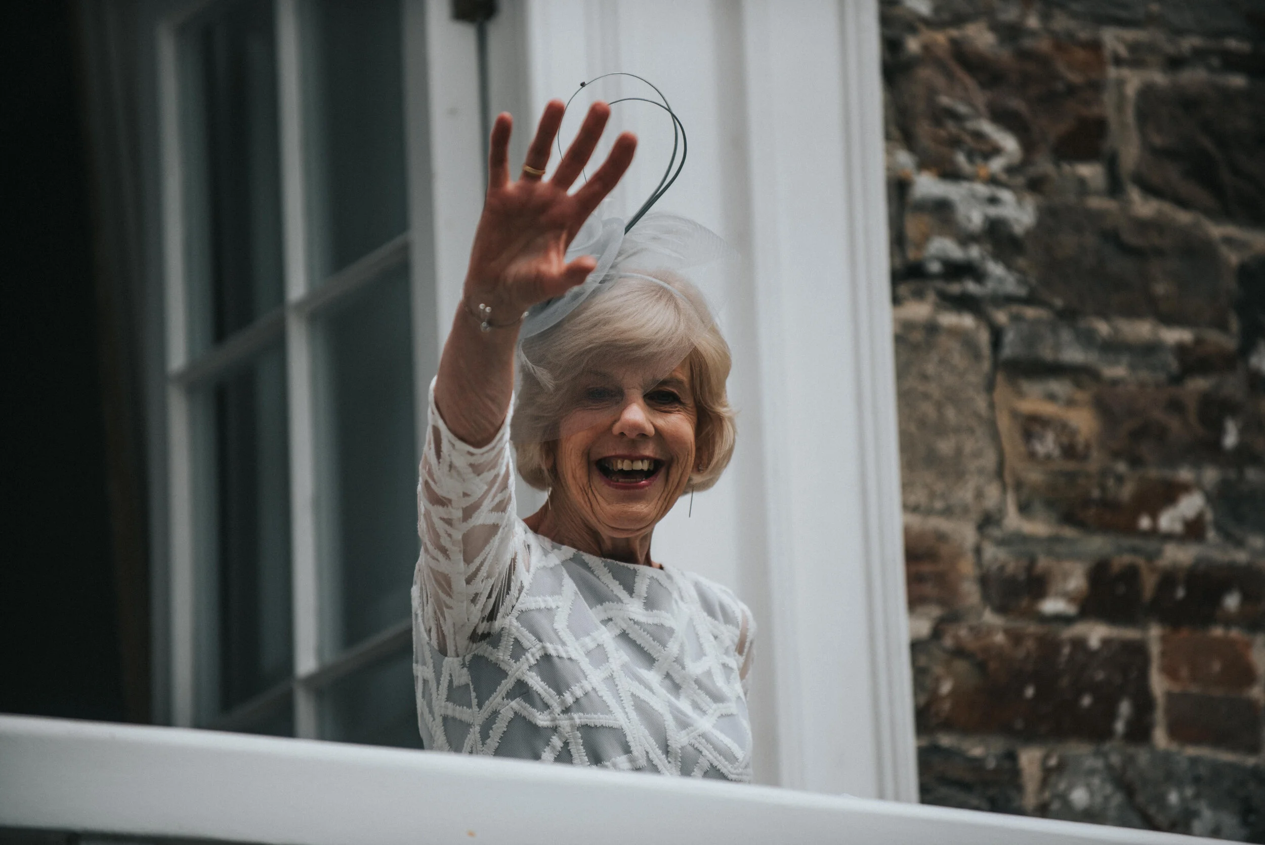 An elderly woman smiling and waving from a balcony, wearing a white lace dress and a fascinator hat - Launcells Barton wedding photographer Cornwall