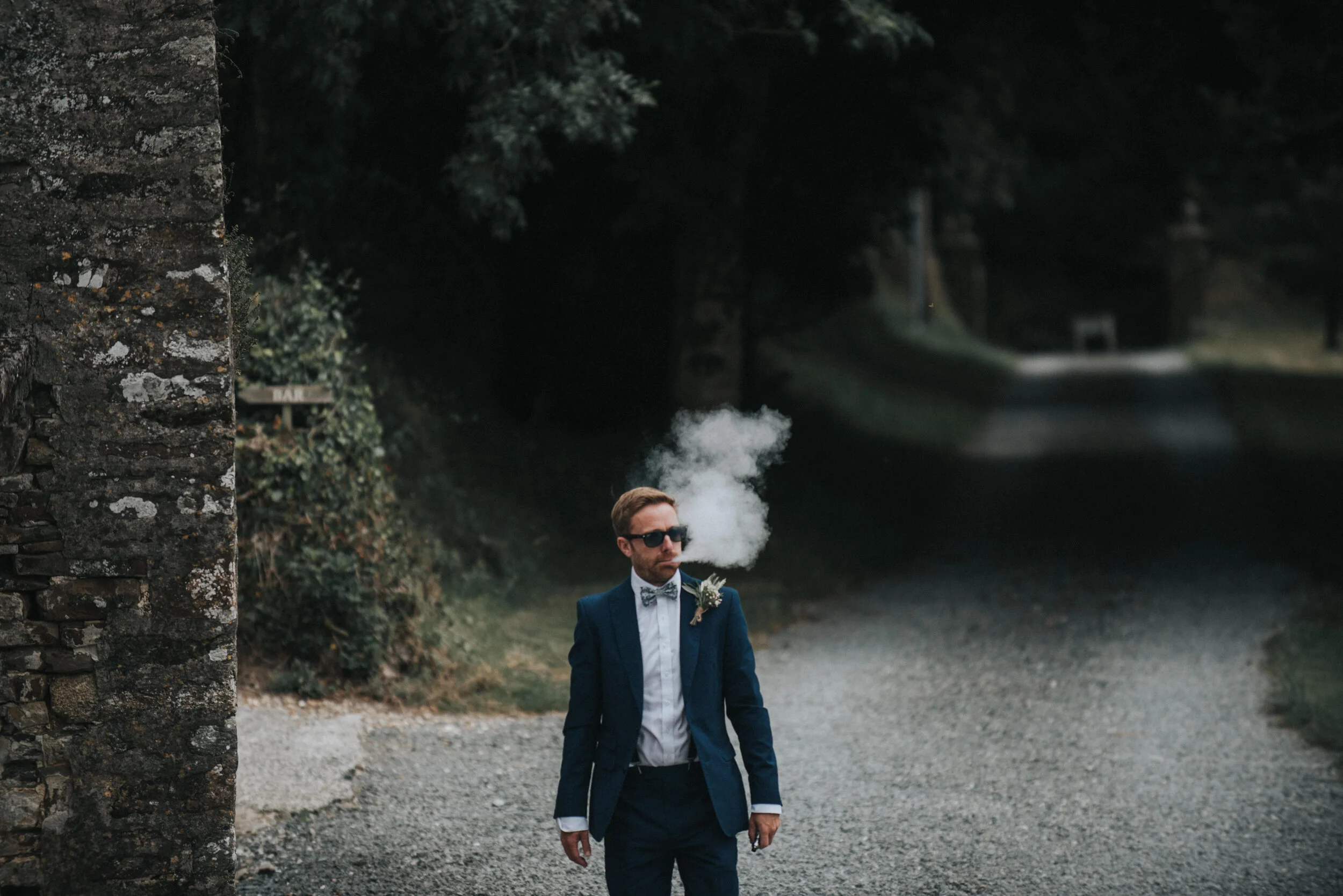 A man in a blue suit, sunglasses, and bow tie walks on a gravel road, smoking with smoke drifting from his mouth, in a rural setting surrounded by trees and a stone wall - Launcells Barton wedding photographer Cornwall