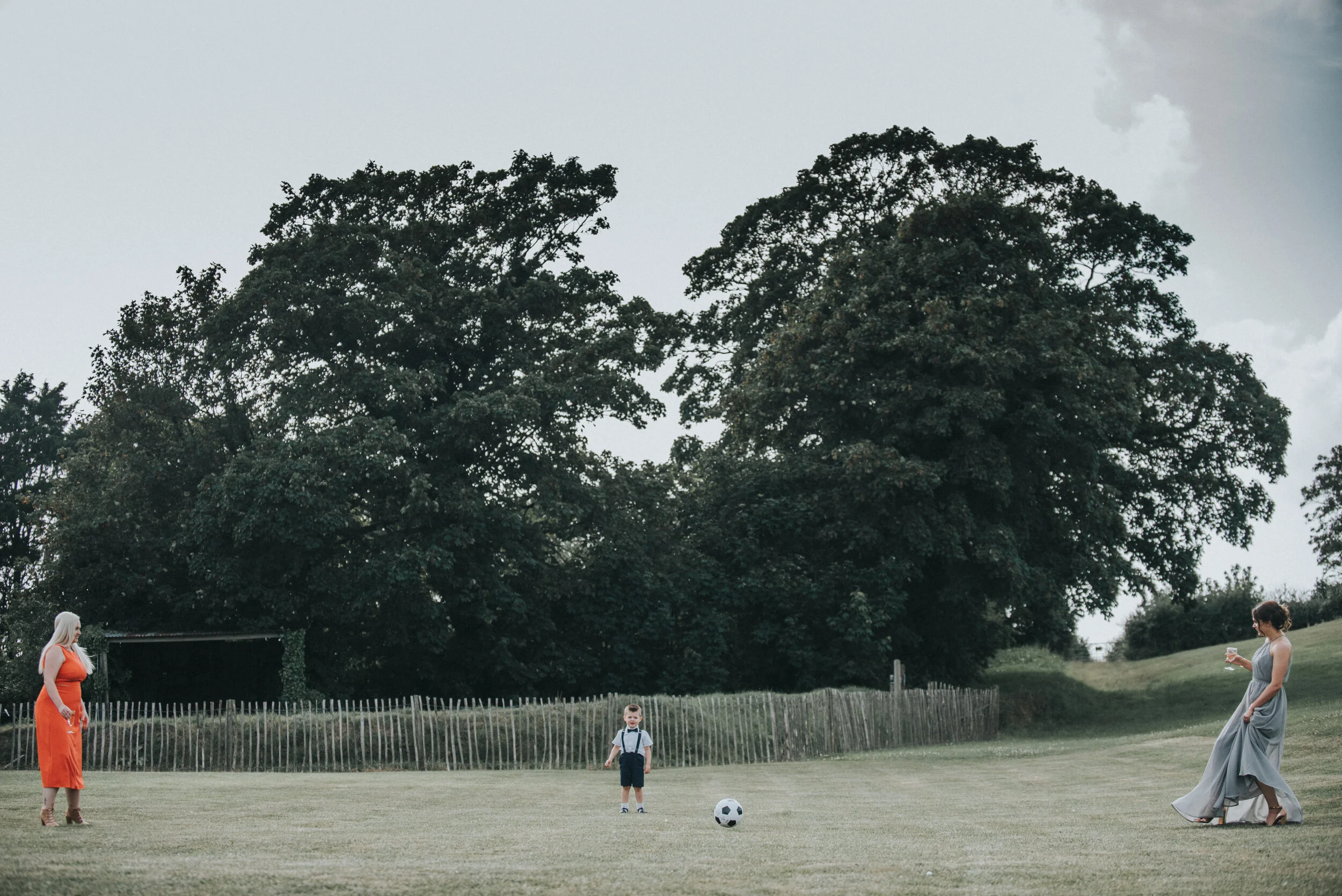 Three people on a grassy field, with two wGuests at Launcells Barton in Bude, captured by Cornwall wedding photographer Mark Shaw Photography


 on opposite sides and a boy in the middle, near a soccer ball, with large trees in the background.