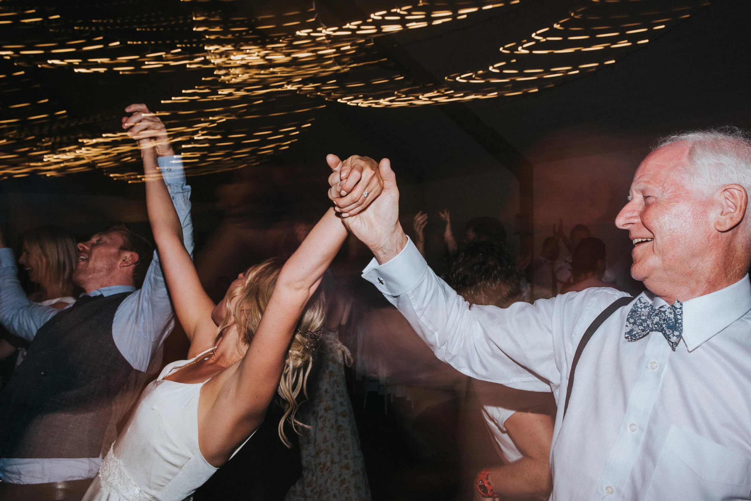 An elderly man in a white dress shirt and bow tie dancing and holding hands with a woman in a white dress at a lively party or wedding reception, with other guests dancing in the background under string lights.