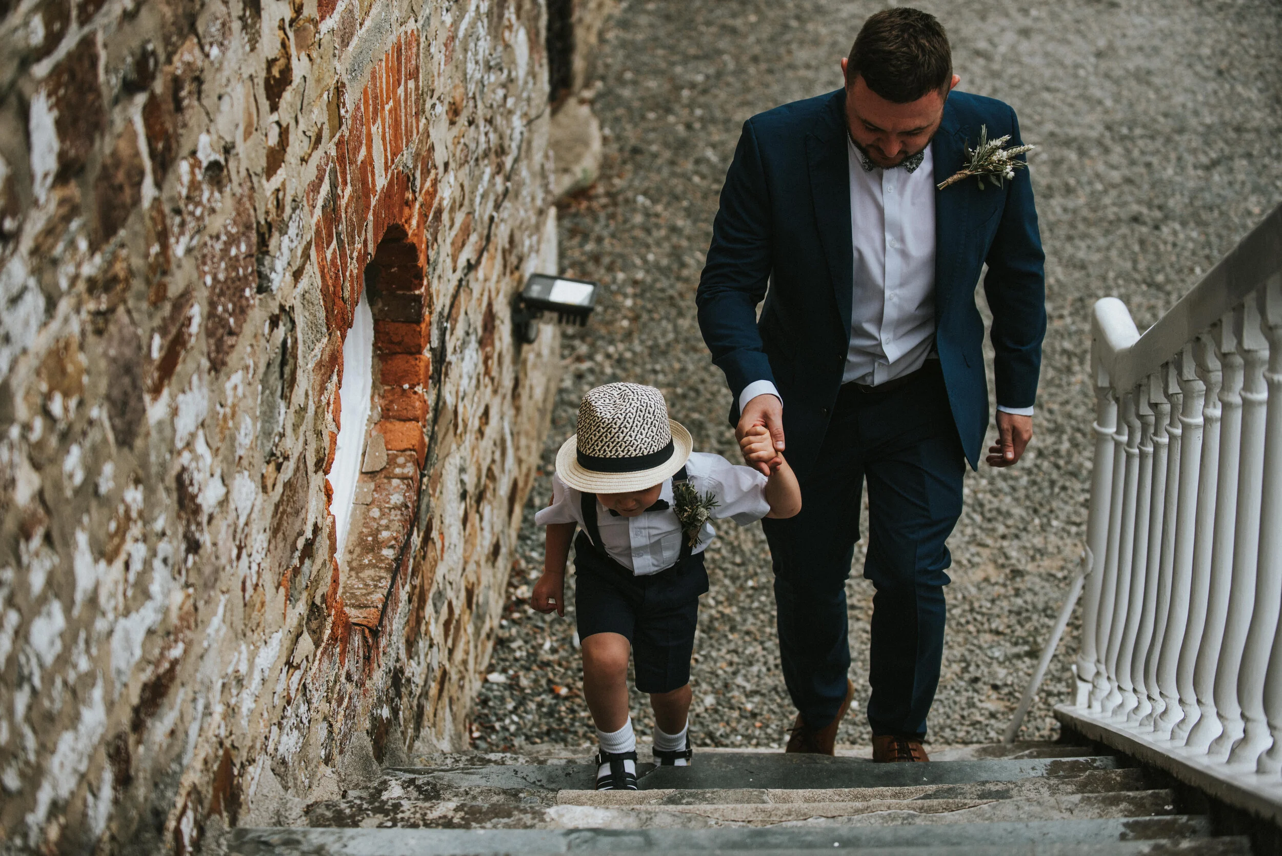 A man in a dark blue suit holding the hand of a young boy dressed in a white shirt, navy shorts, black and white shoes, and a straw hat as they ascend stone steps outside a brick building - Launcells Barton wedding photographer Cornwall