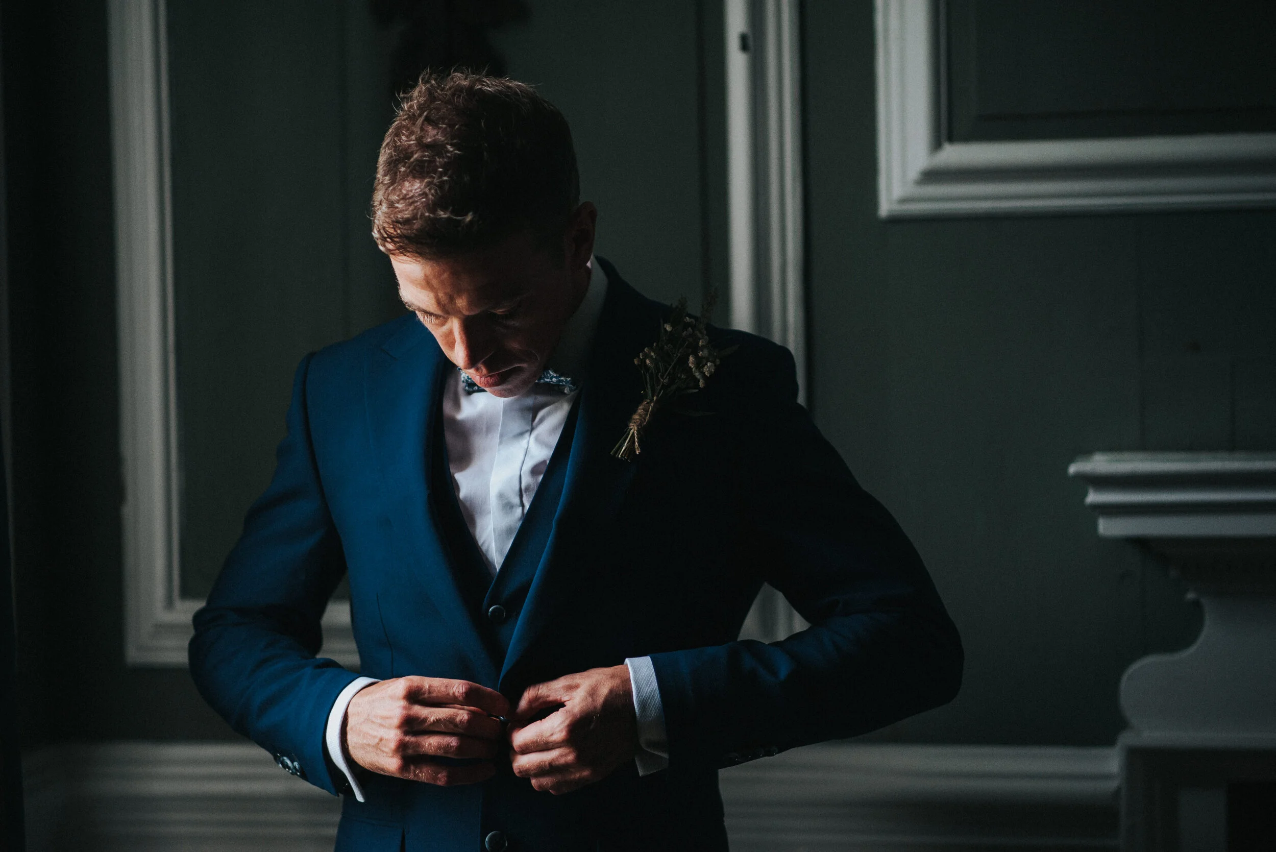 A man in a dark blue suit, white shirt, and bow tie is adjusting his suit button, standing indoors near a window with dark walls and white trim, with a boutonniere pinned to his lapel - Launcells Barton wedding photographer Cornwall