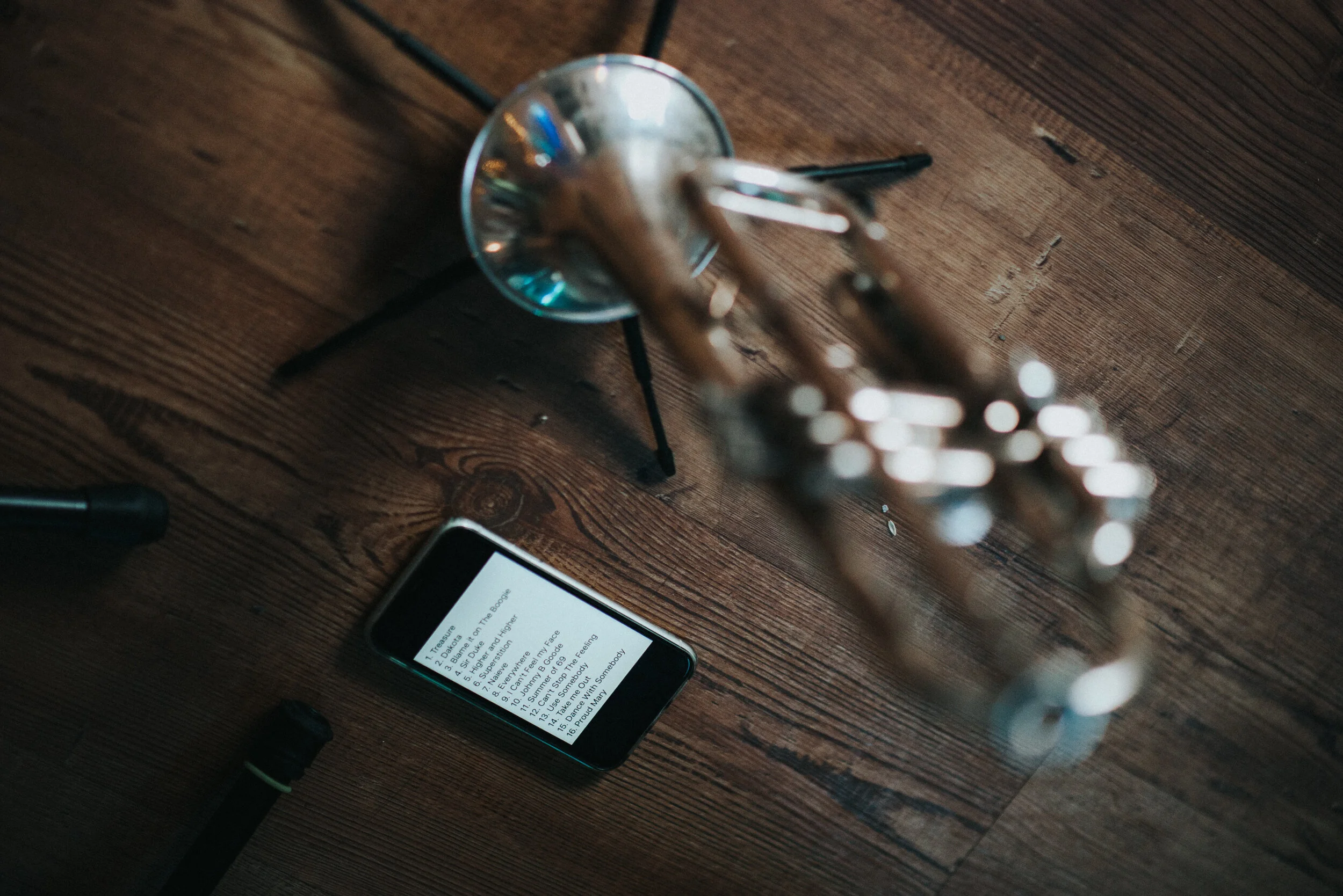 A wooden table holds a smartphone displaying song lyrics, a pen, and a brass trumpet in a metal stand.
