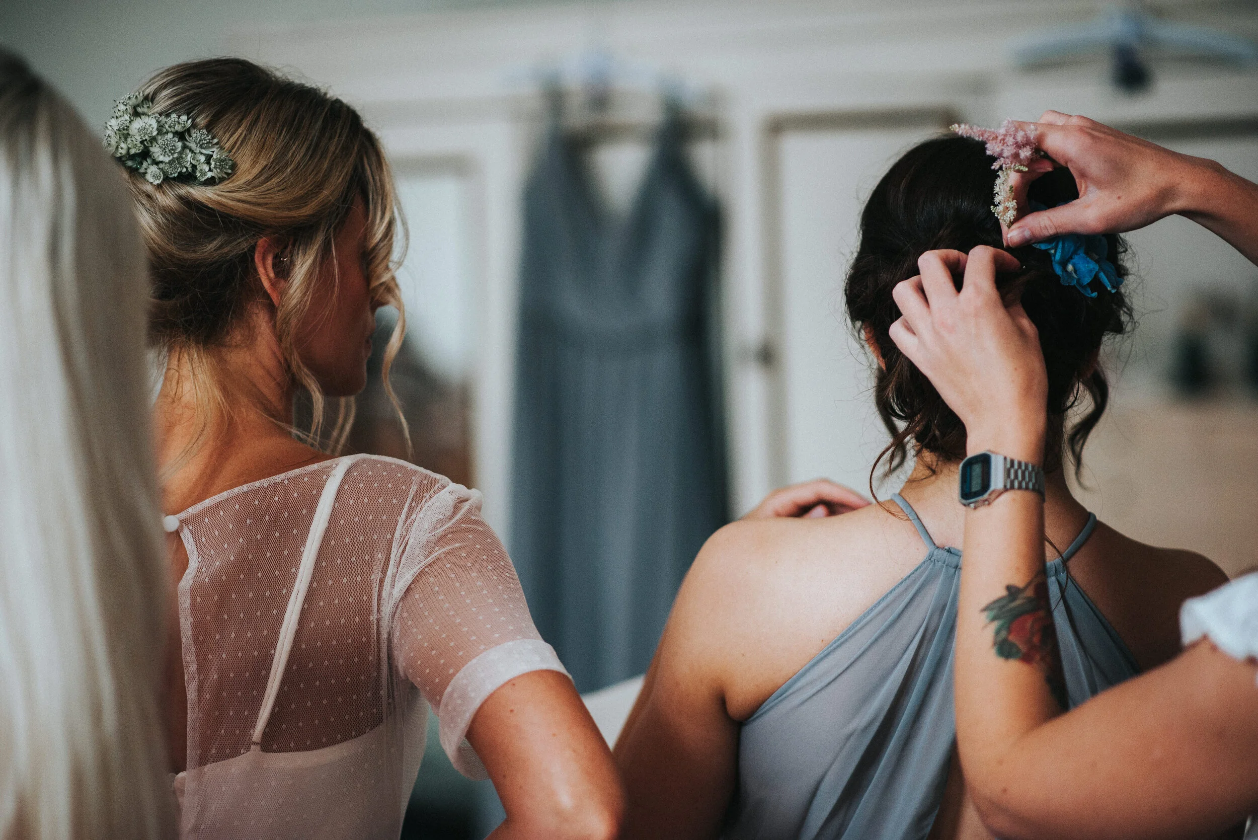 A woman is having her hair styled with flowers, while another woman assists her, in a room with a dress hanging in the background - Launcells Barton wedding photographer Cornwall
