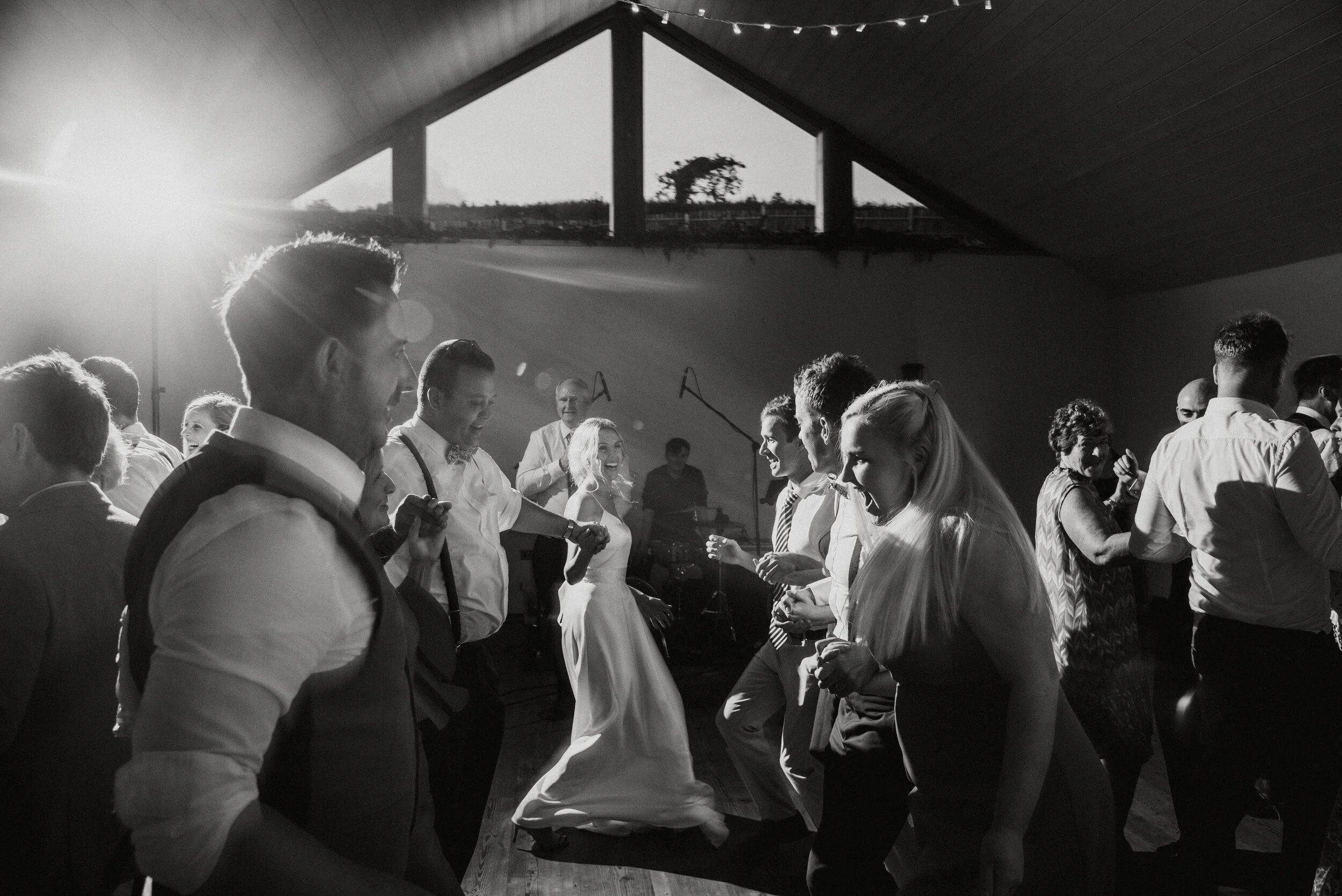 People dancing at a wedding reception inside a venue with a triangular ceiling and large window, sunlight streaming in from outside.