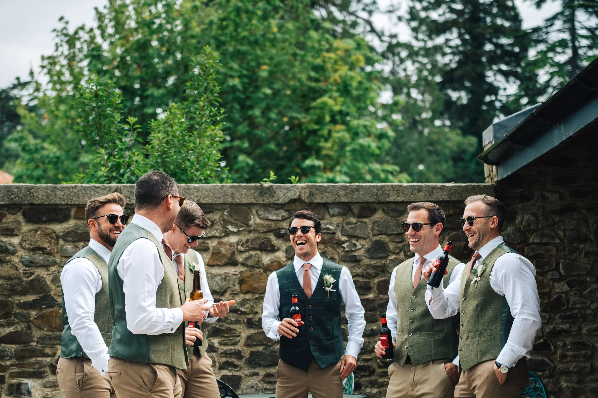 Groomsmen in vests laughing and holding drinks outdoors at a wedding.