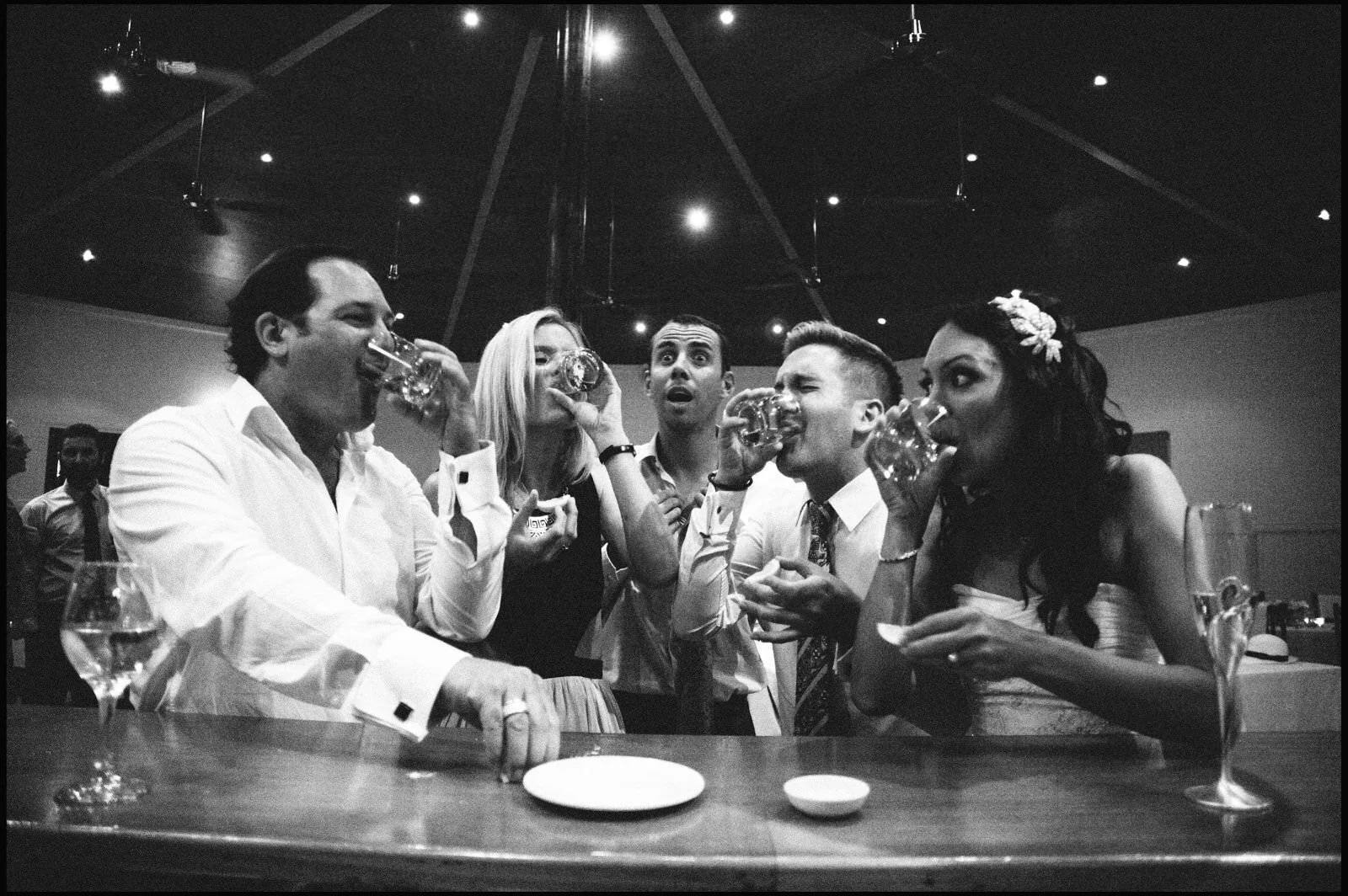A group of five friends at a party, drinking drinks from glasses, standing around a bar, in a room with a high ceiling and dim lighting.