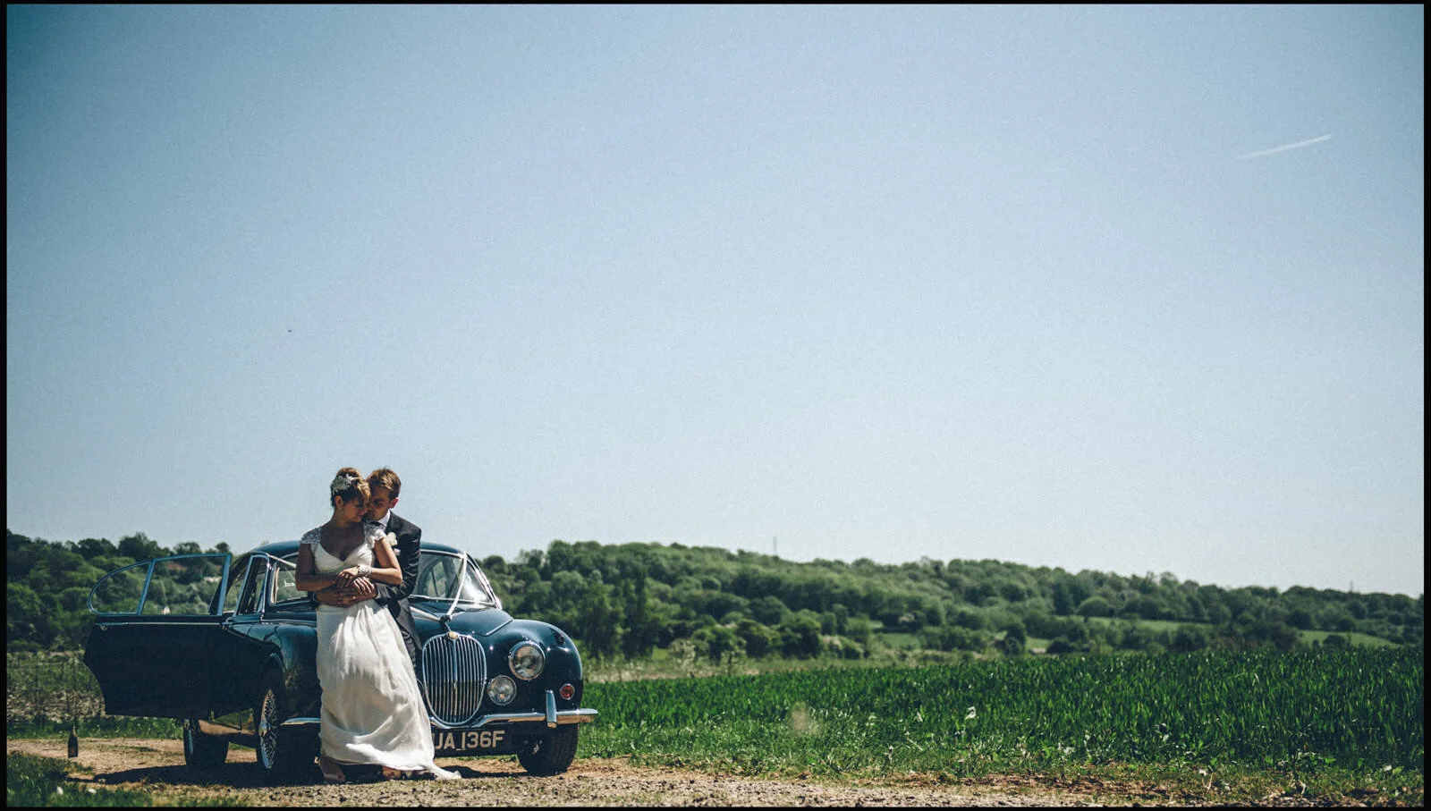 A bride and groom in wedding attire standing close together beside a vintage black car in a rural field with green hills in the background under a clear blue sky.