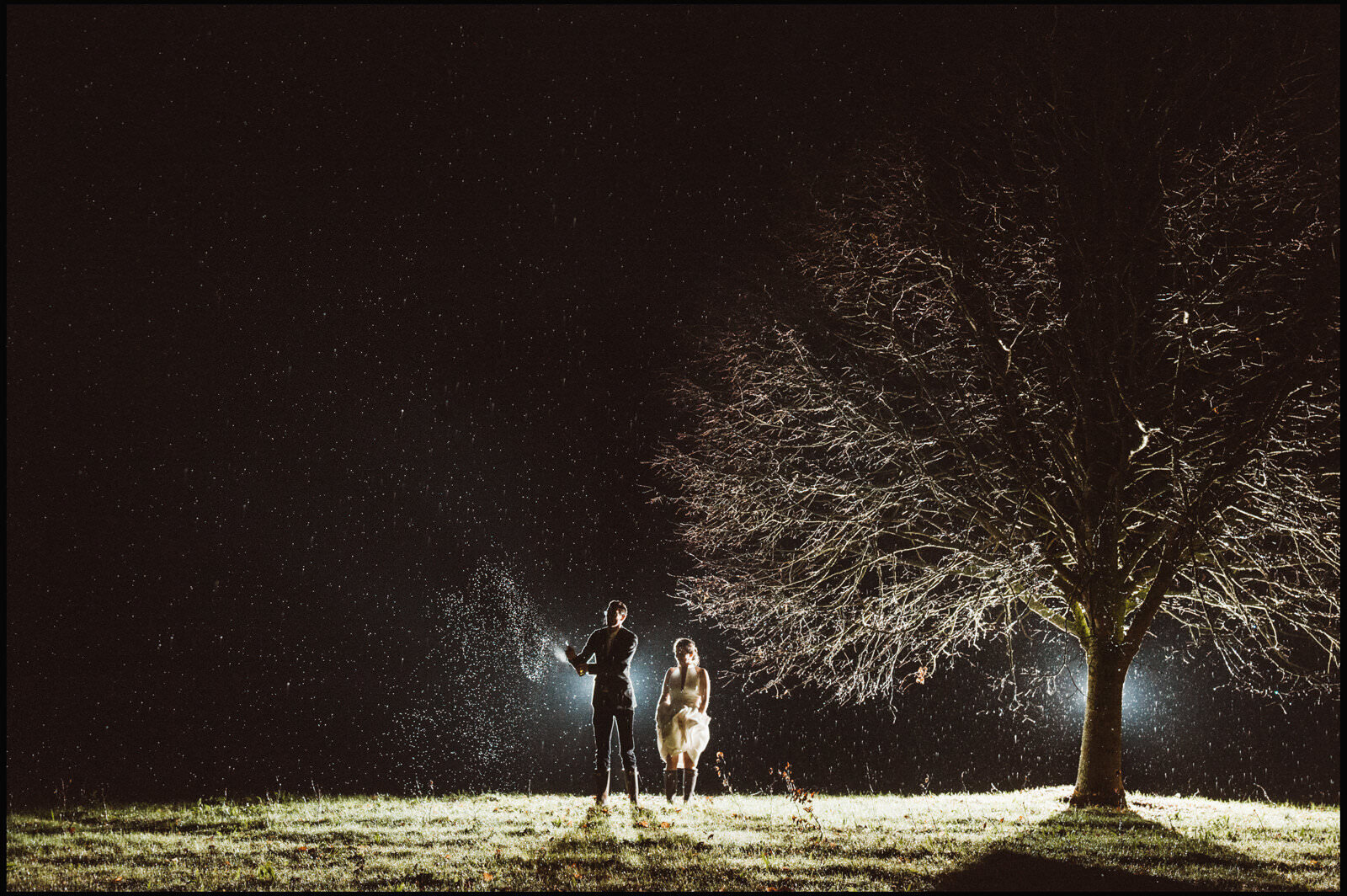 A man and woman celebrating New Year's Eve at night outdoors, with the man holding a party popper and confetti in the air, next to a large tree under a starry sky.