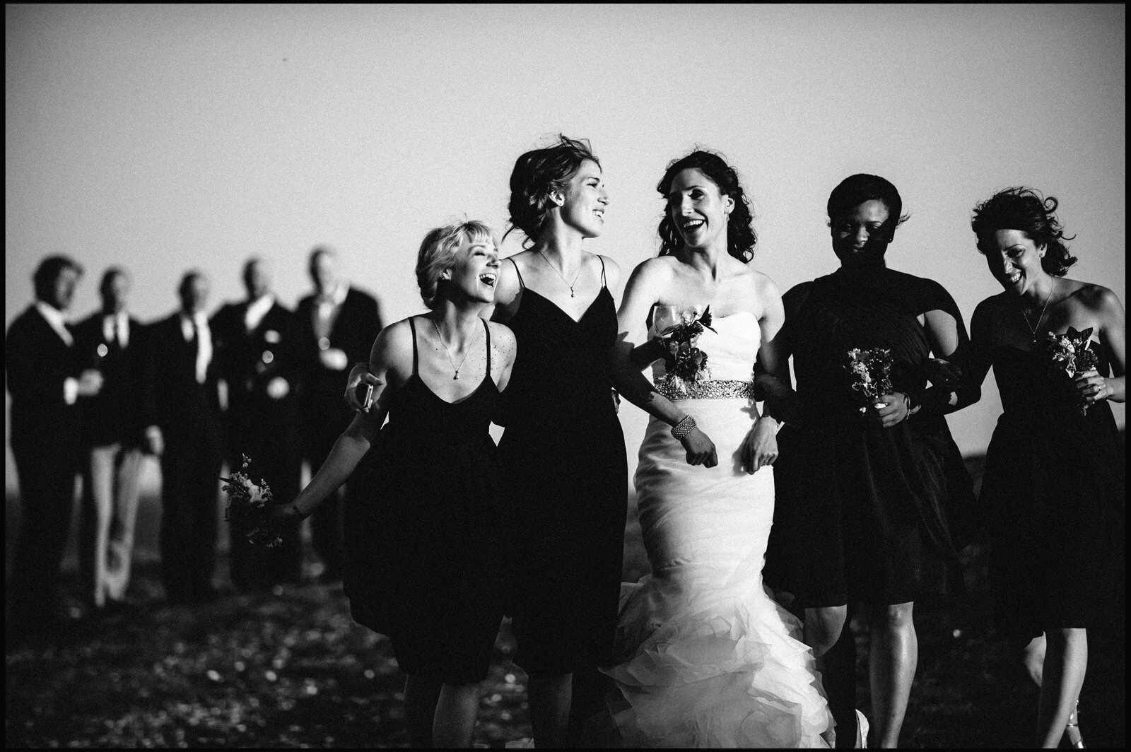 Black and white photo of a bride and five women walking together, smiling and holding bouquets, with a line of men in suits blurred in the background.