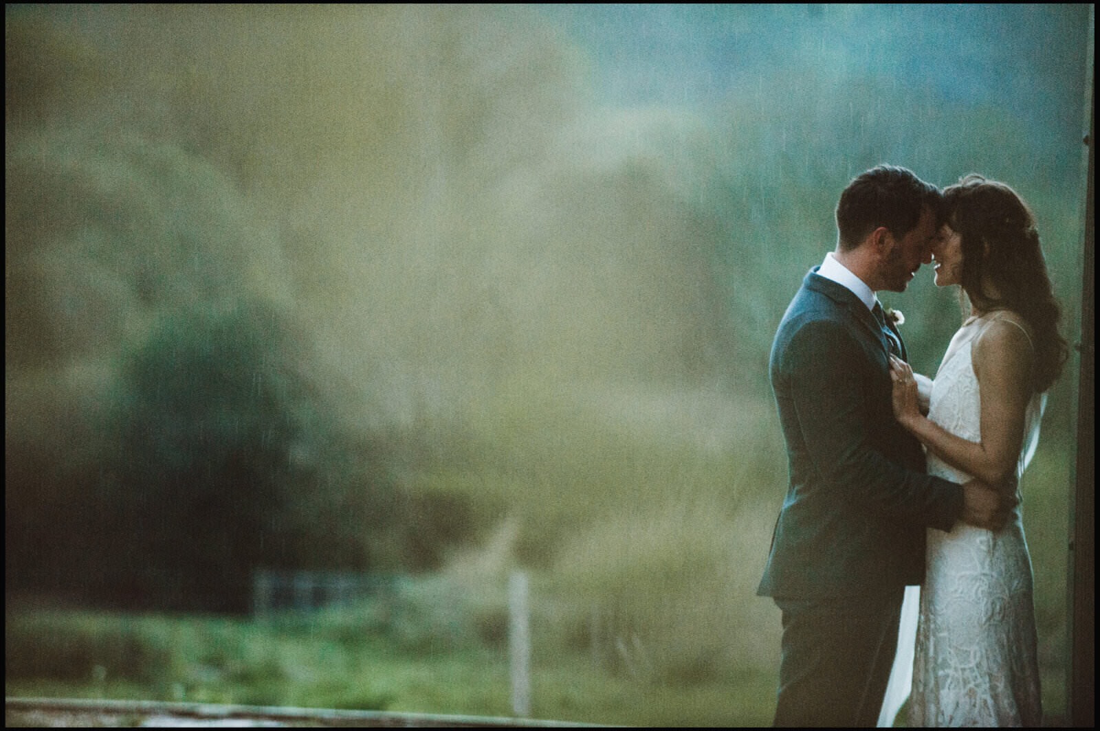 A bride and groom standing close, touching foreheads, in an outdoor setting during a rainstorm, with the groom in a dark suit and the bride in a white lace dress.