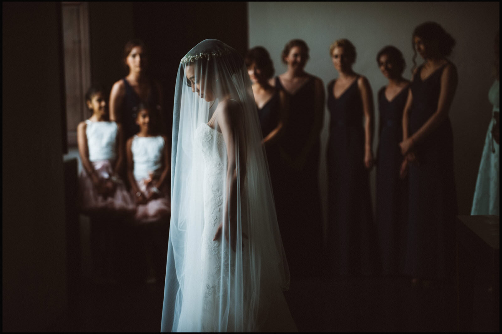 A bride in a white wedding dress and veil stands with her head bowed, surrounded by women in black dresses and two young girls in white tops and pink skirts, in a dimly lit room.
