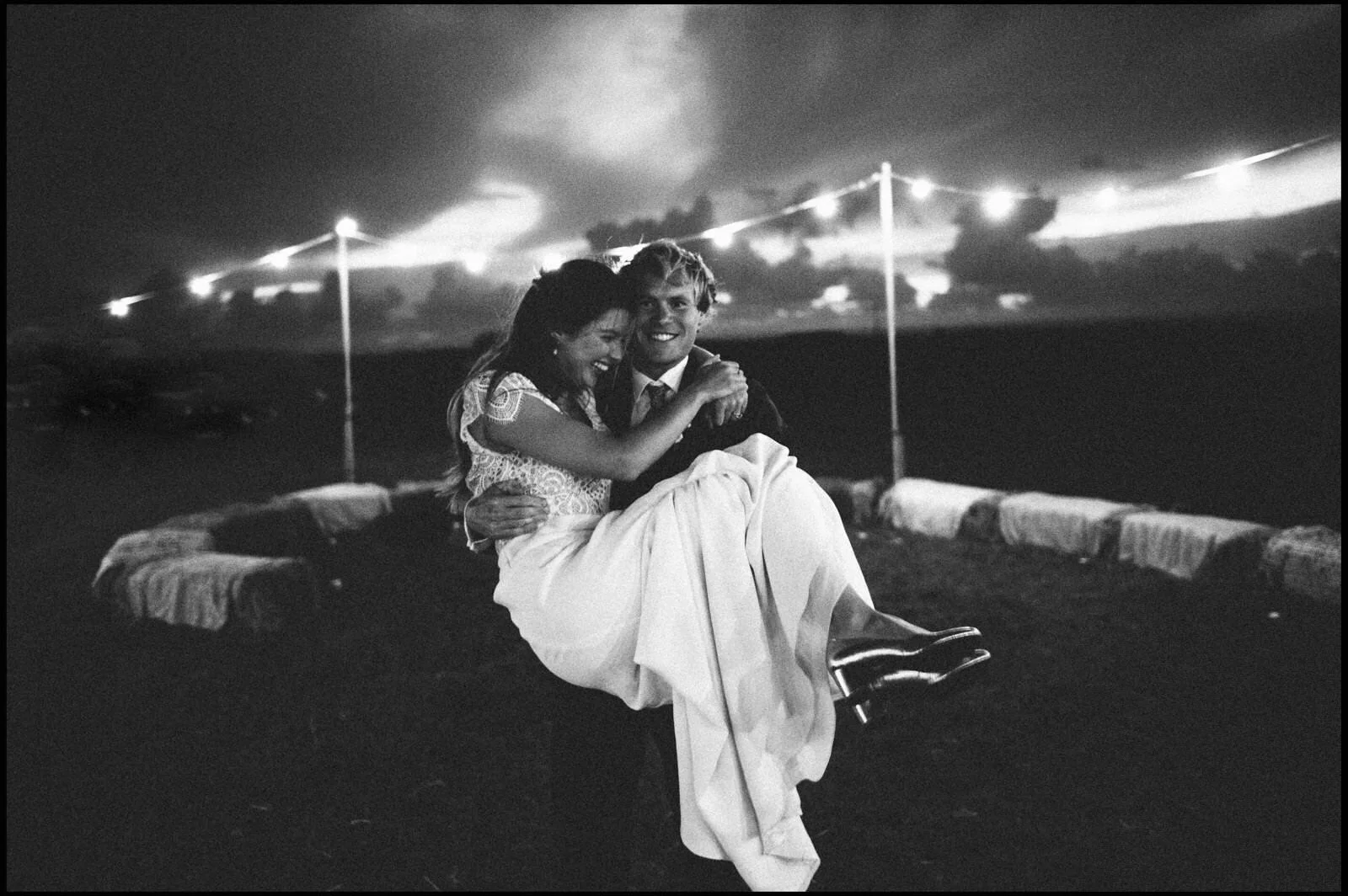 Black and white photo of a couple sitting together on a swing at night, smiling and embracing, with fireworks in the sky behind them.